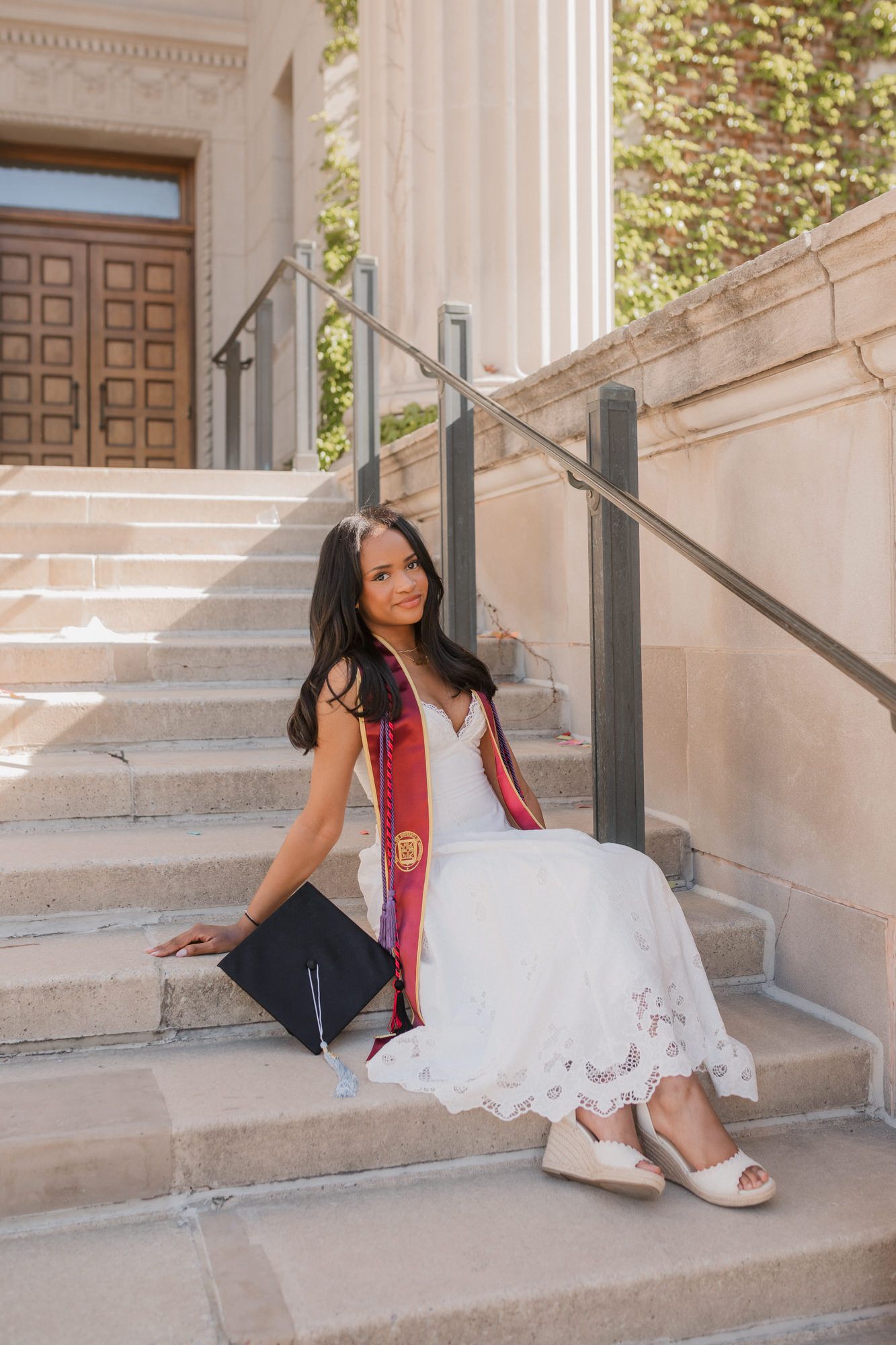 Young woman in white lace dress with graduation sash and cap sits on stone steps of university building.