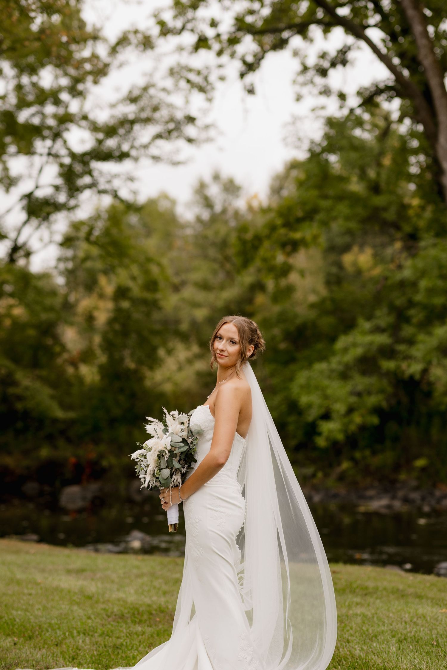 Bride in a white fitted gown and long veil holds a bouquet, posing outdoors with lush green trees behind her.