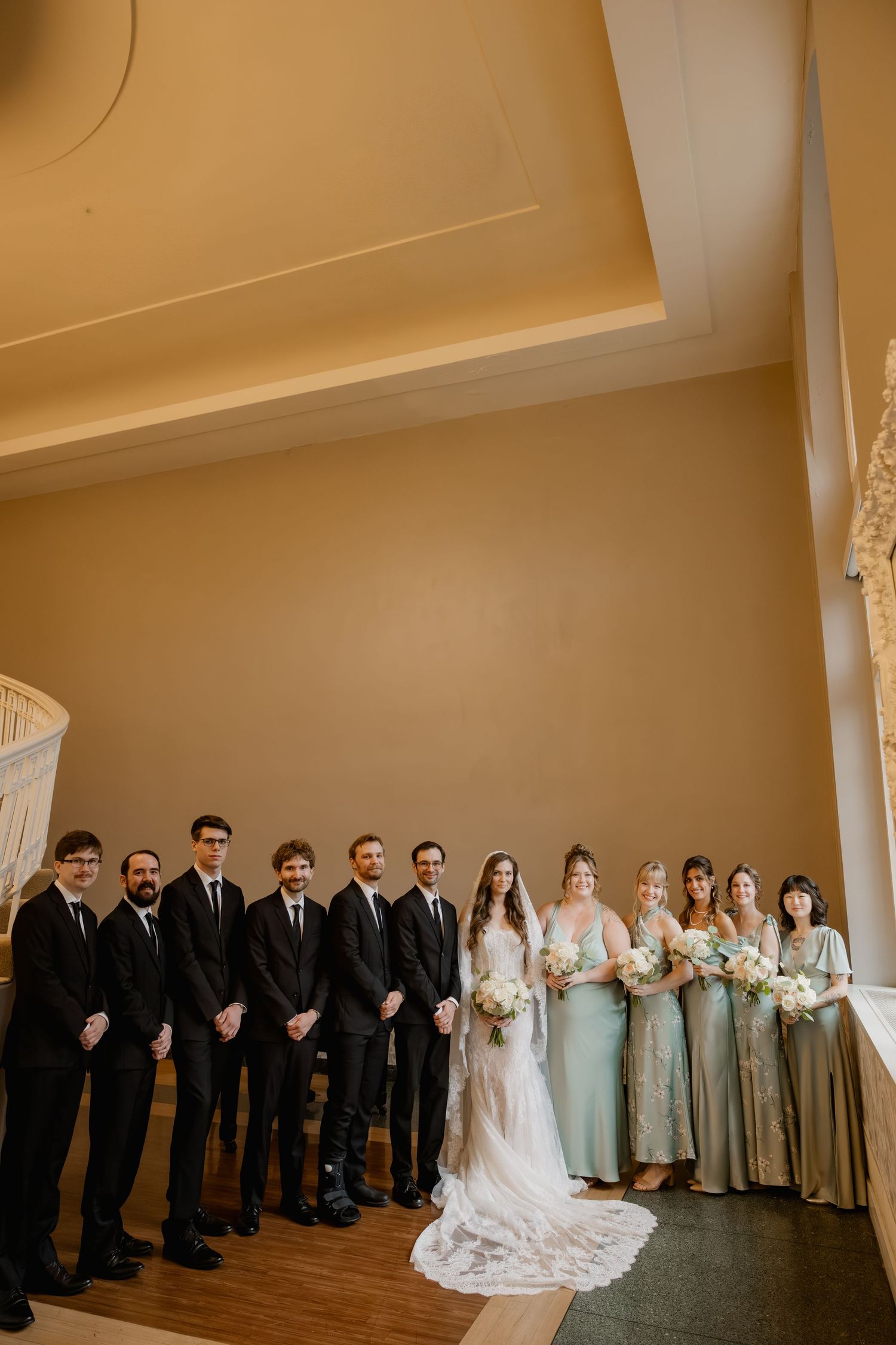 Wedding party poses indoors, groomsmen in black suits beside bride in lace gown and bridesmaids in sage green dresses.