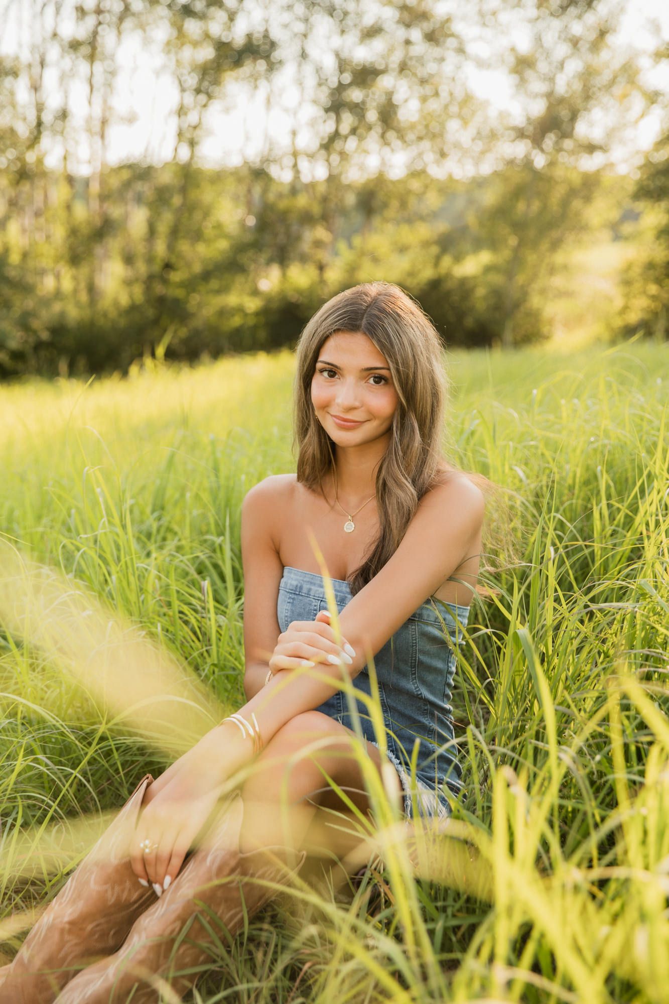 Young woman in denim outfit sitting in lush green meadow grass, smiling at camera during golden hour outdoor photo session.