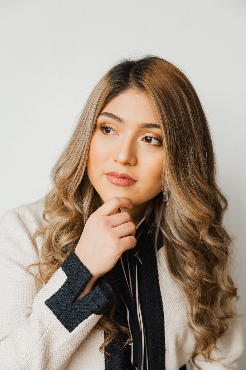 Young woman with wavy blonde hair in white blazer, posing thoughtfully with hand on chin against light background.