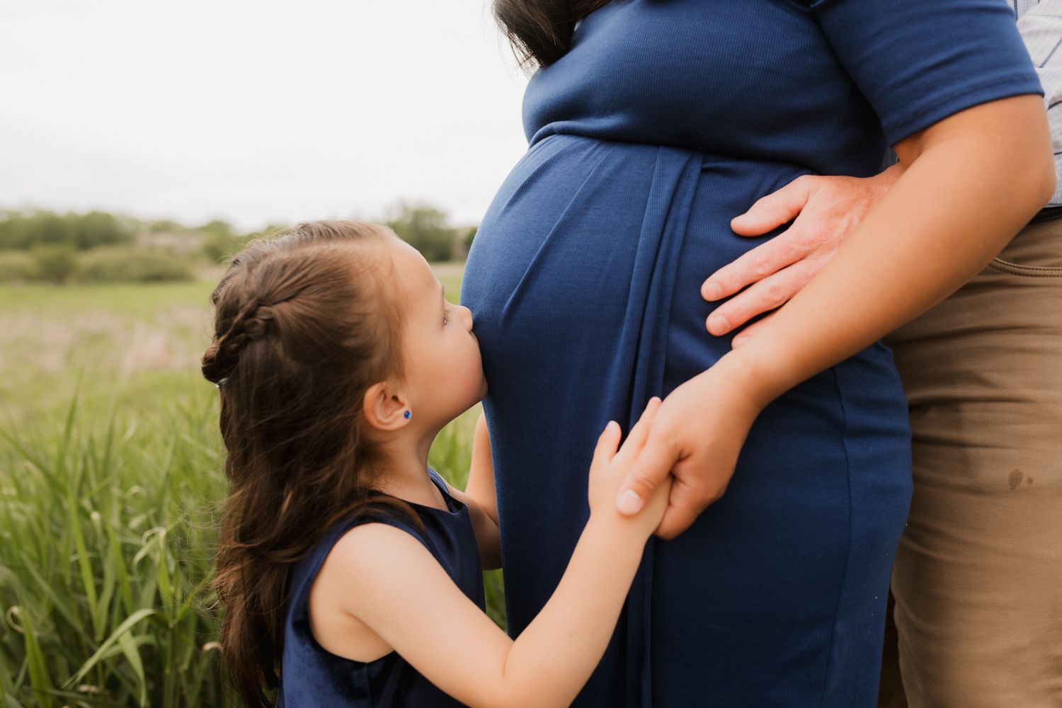 A young girl kisses a pregnant woman's belly outdoors in a green field during a maternity photo session.