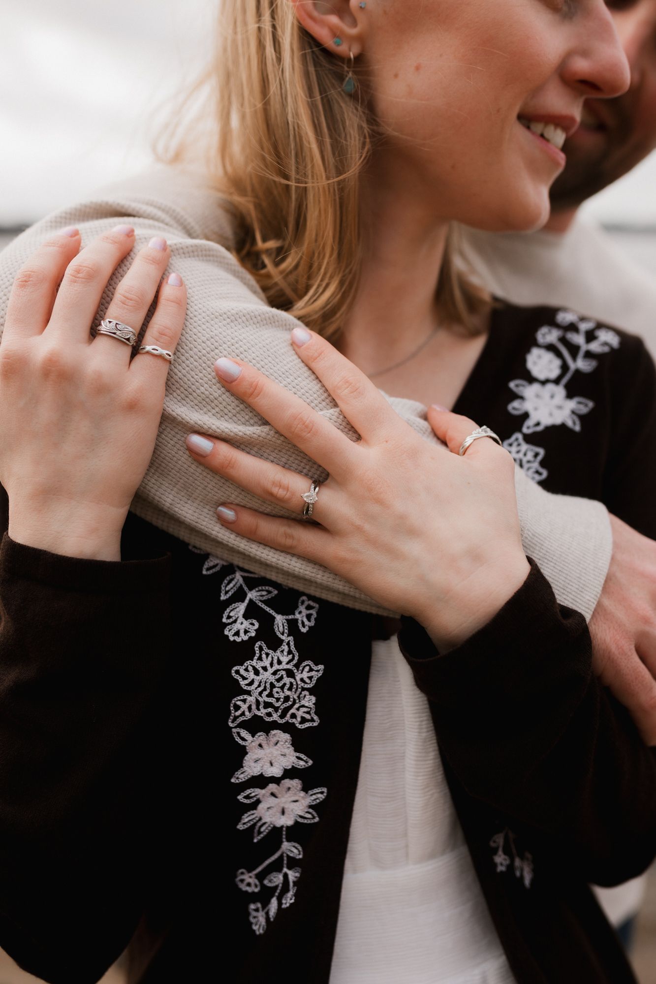 Close-up of a couple embracing, showcasing elegant rings on the woman's hands and floral embroidered black jacket.