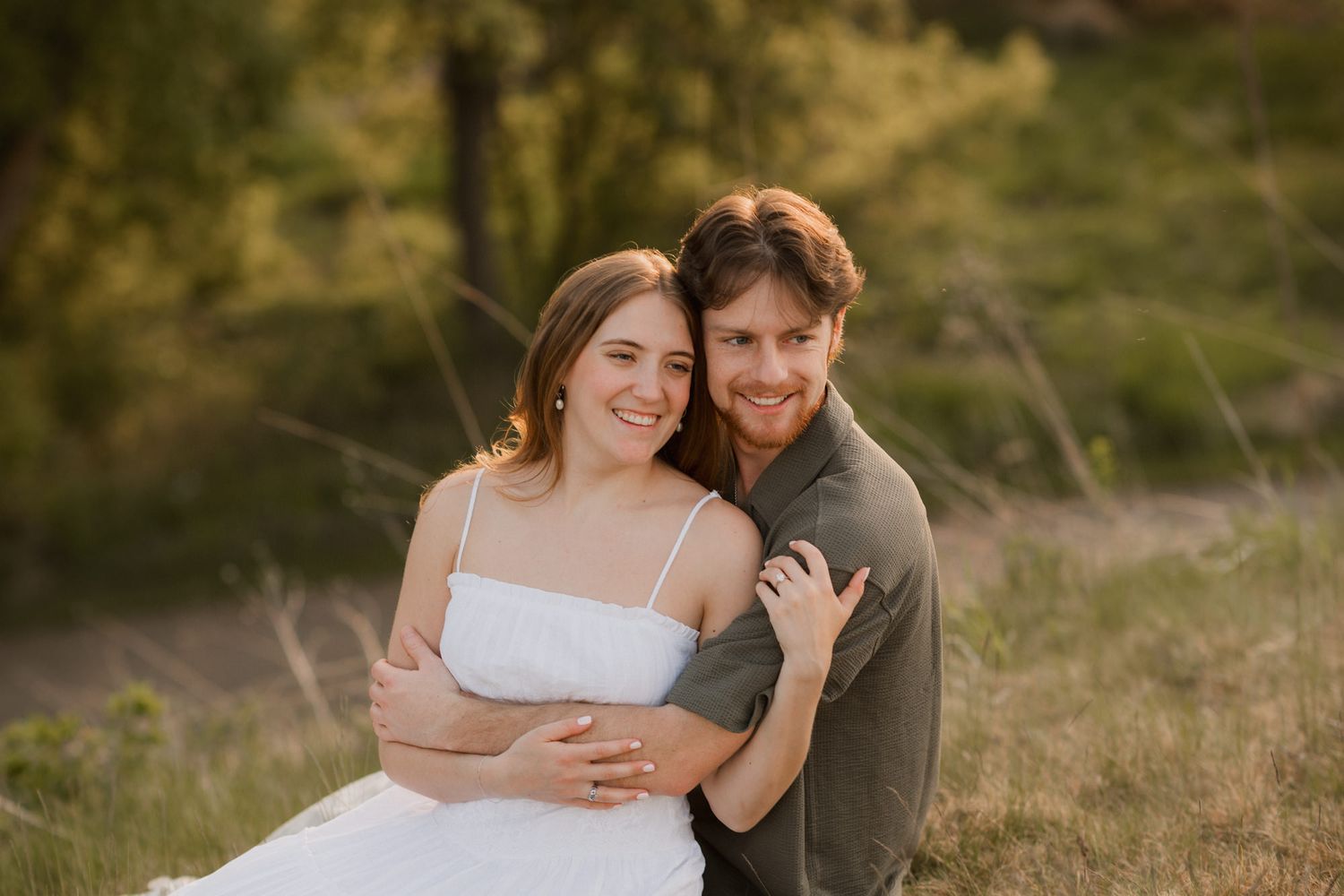 A smiling couple poses together in a sun-kissed field, the man embracing the woman from behind.