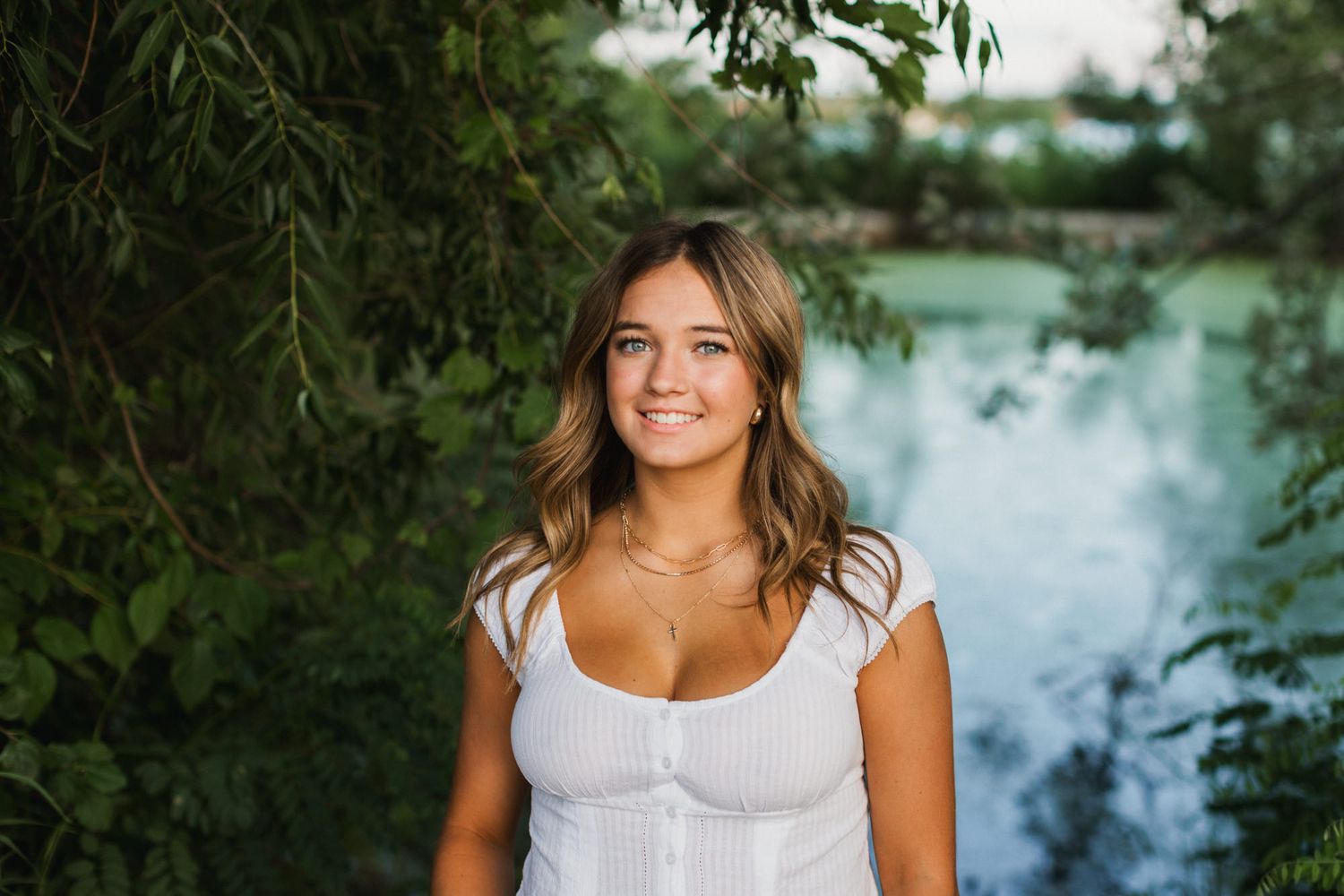 Young woman in white dress smiling outdoors near a serene lake surrounded by lush green trees.