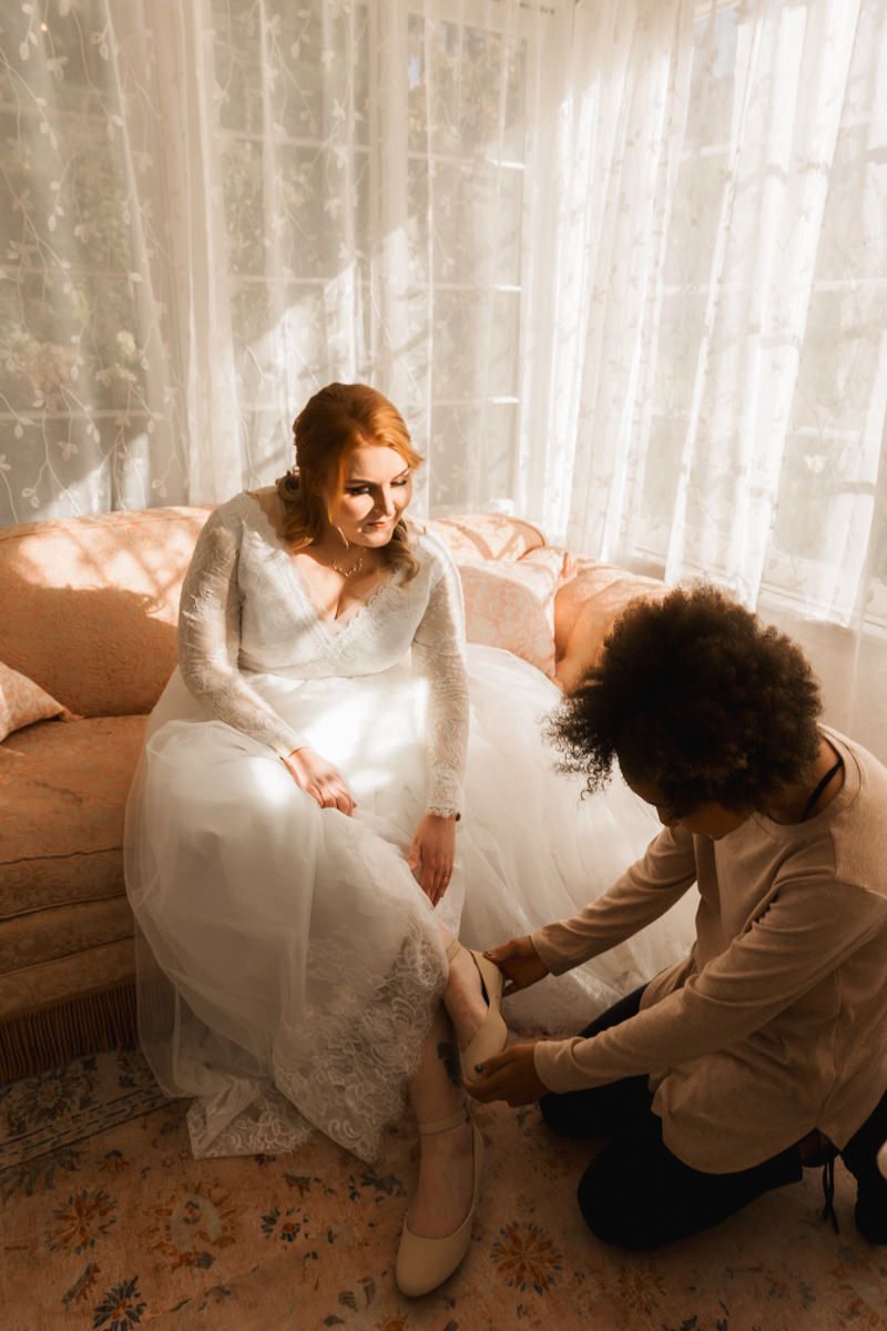 A bride in a white lace gown sits on a couch while getting help with her shoes in a sunlit room with sheer curtains.