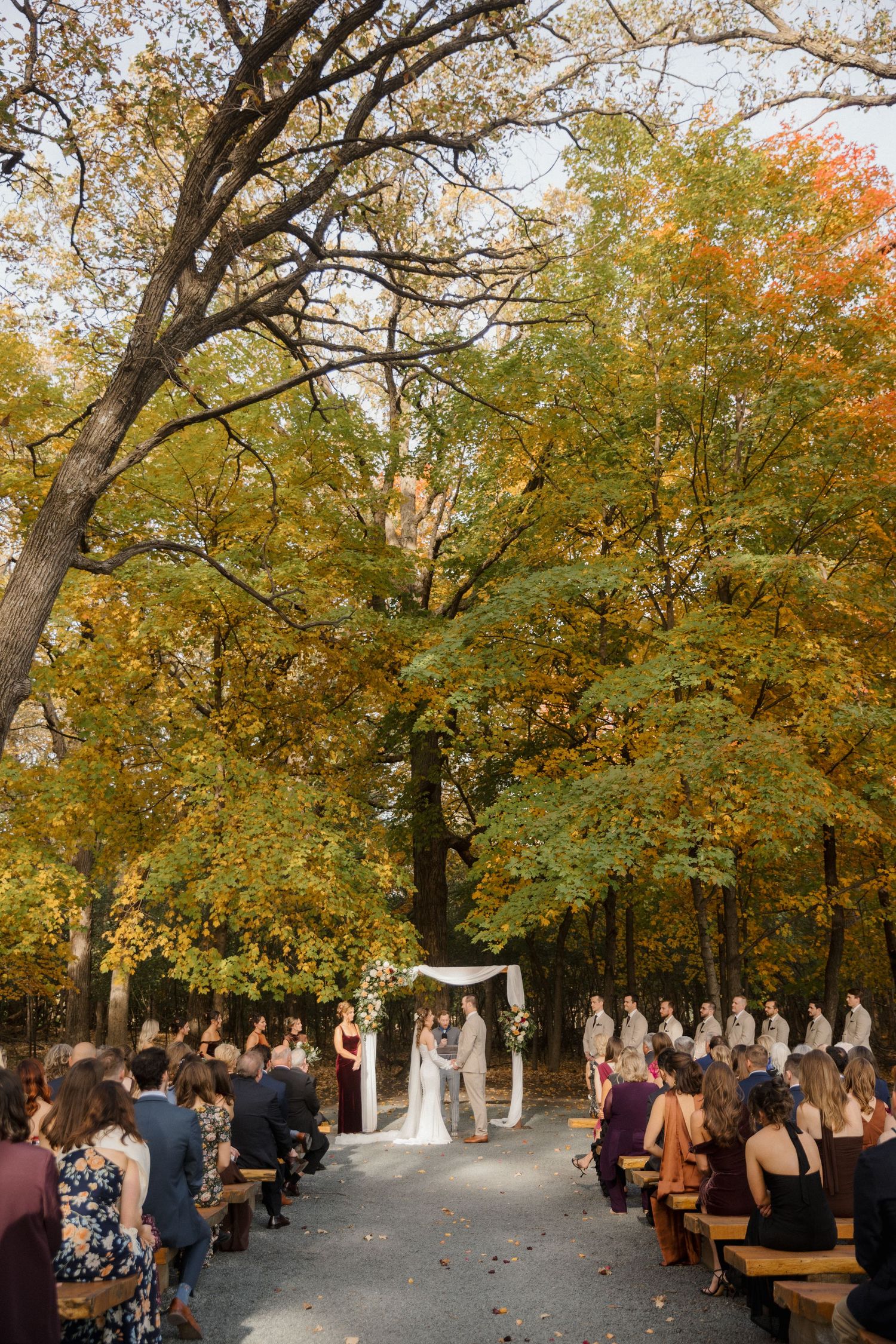 Couple exchanges vows beneath a floral arch as guests watch from wooden benches among colorful autumn trees.