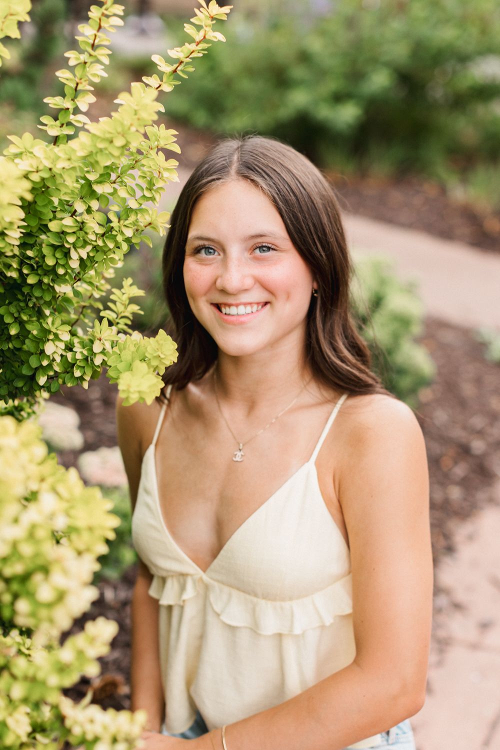 A person in a cream-colored spaghetti strap top poses among yellow-green foliage in an outdoor setting.