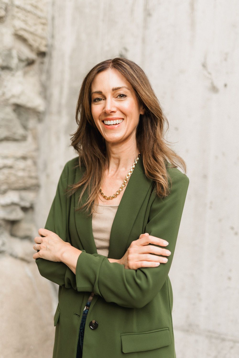 Smiling woman in green blazer and gold necklace with arms crossed, posing confidently against a stone wall.