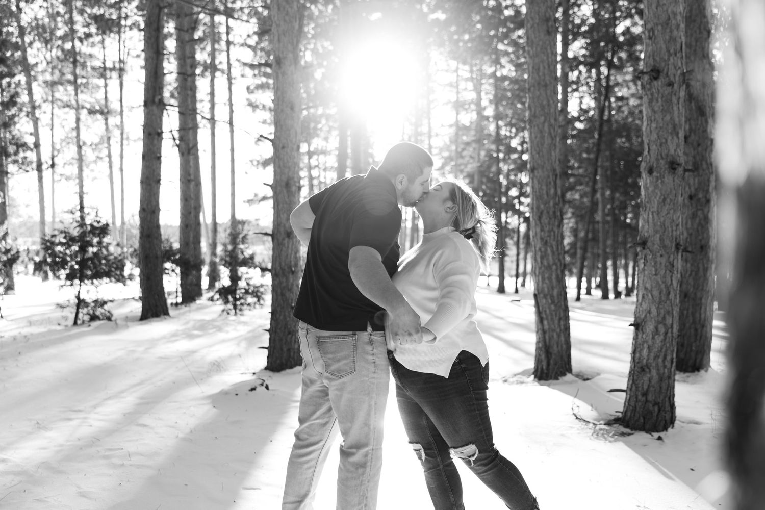 Black and white photo of couple kissing in snowy pine forest with sunlight streaming through trees.