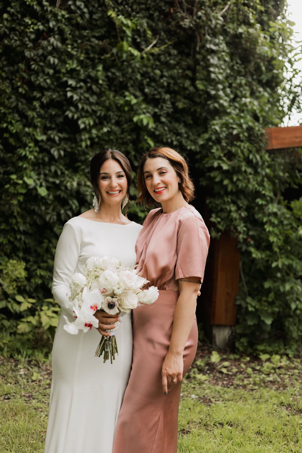 Two people in white and pink dresses share a joyful moment together in a garden setting with lush greenery.