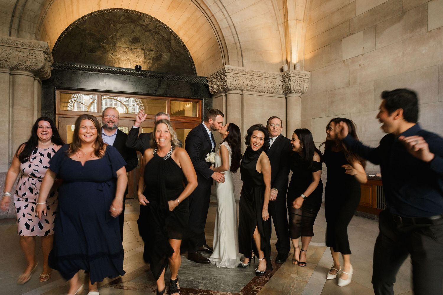 Wedding party celebrating in elegant stone archway venue, bride in white gown surrounded by guests in black attire.