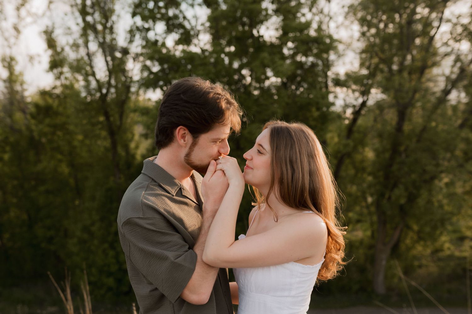 A romantic couple shares a tender moment outdoors, the man kissing the woman's hand among lush green trees.