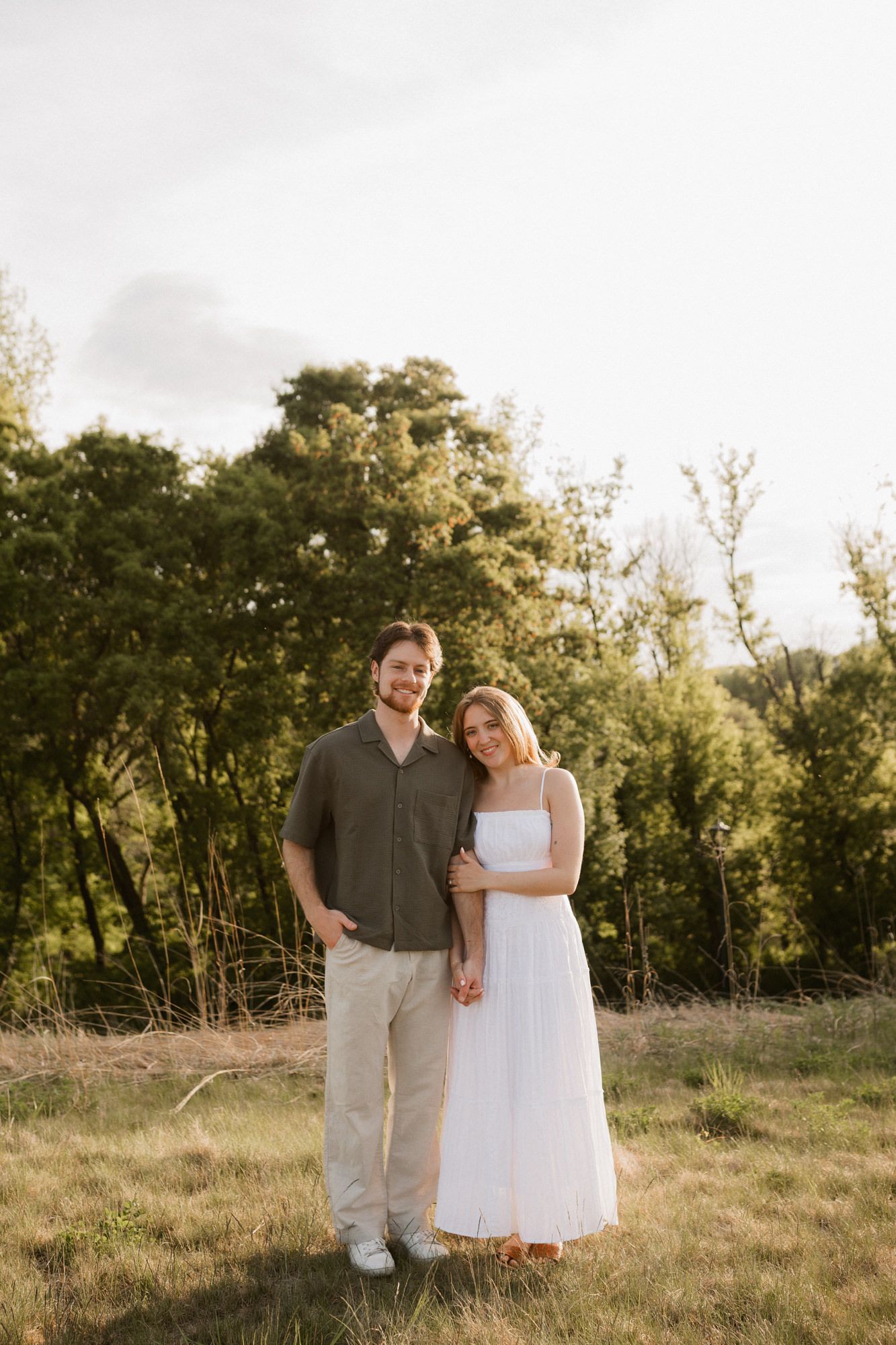 Couple posing outdoors in golden hour light, man in olive shirt and woman in white dress, surrounded by lush green trees.