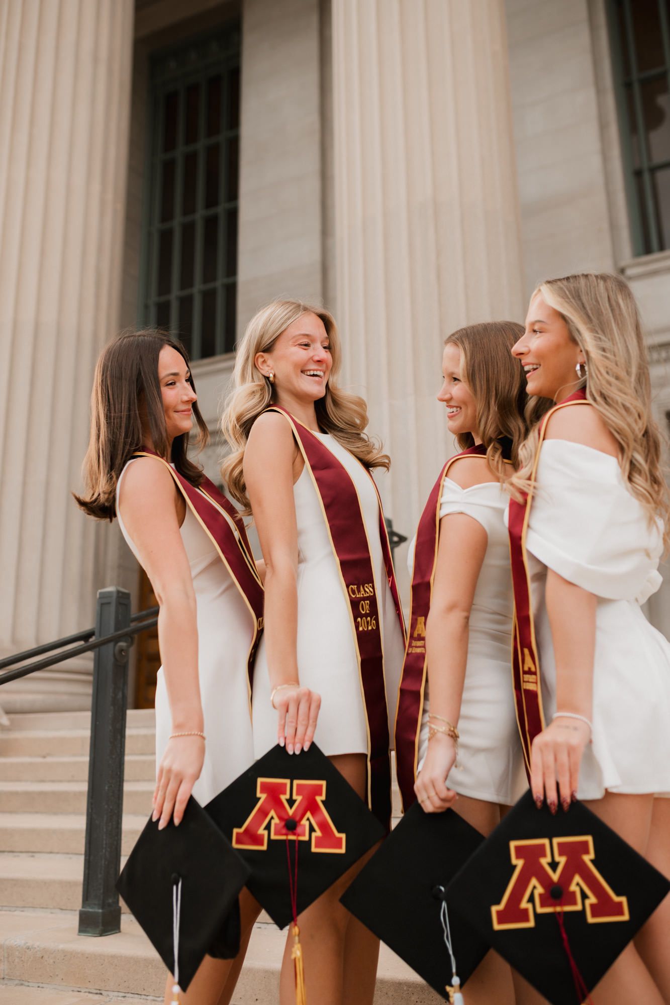Four University of Minnesota graduates in white dresses holding graduation caps with the Gopher M logo on steps.