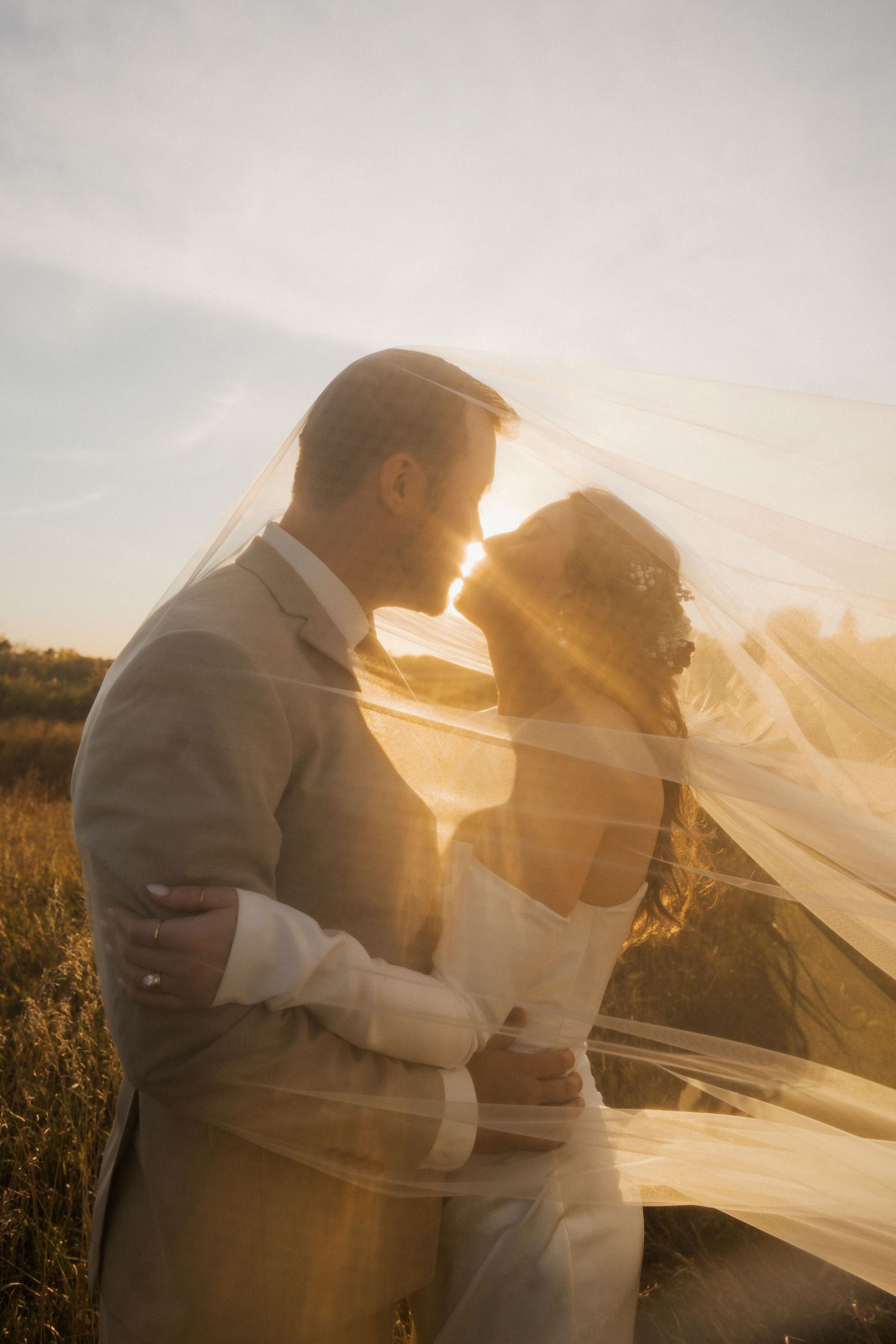 Bride and groom embrace in a golden sunset field as a sheer veil drifts over them, sunlight glowing between their faces.