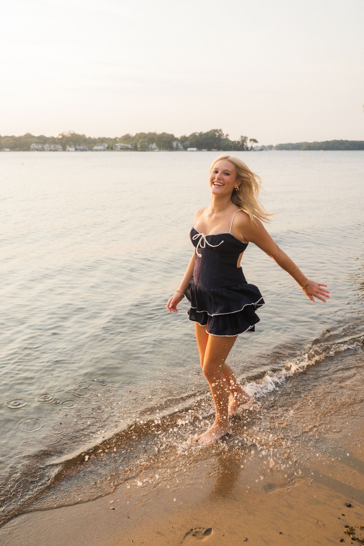 Smiling blonde woman in black dress running barefoot along sandy beach shore with water splashing around her feet.