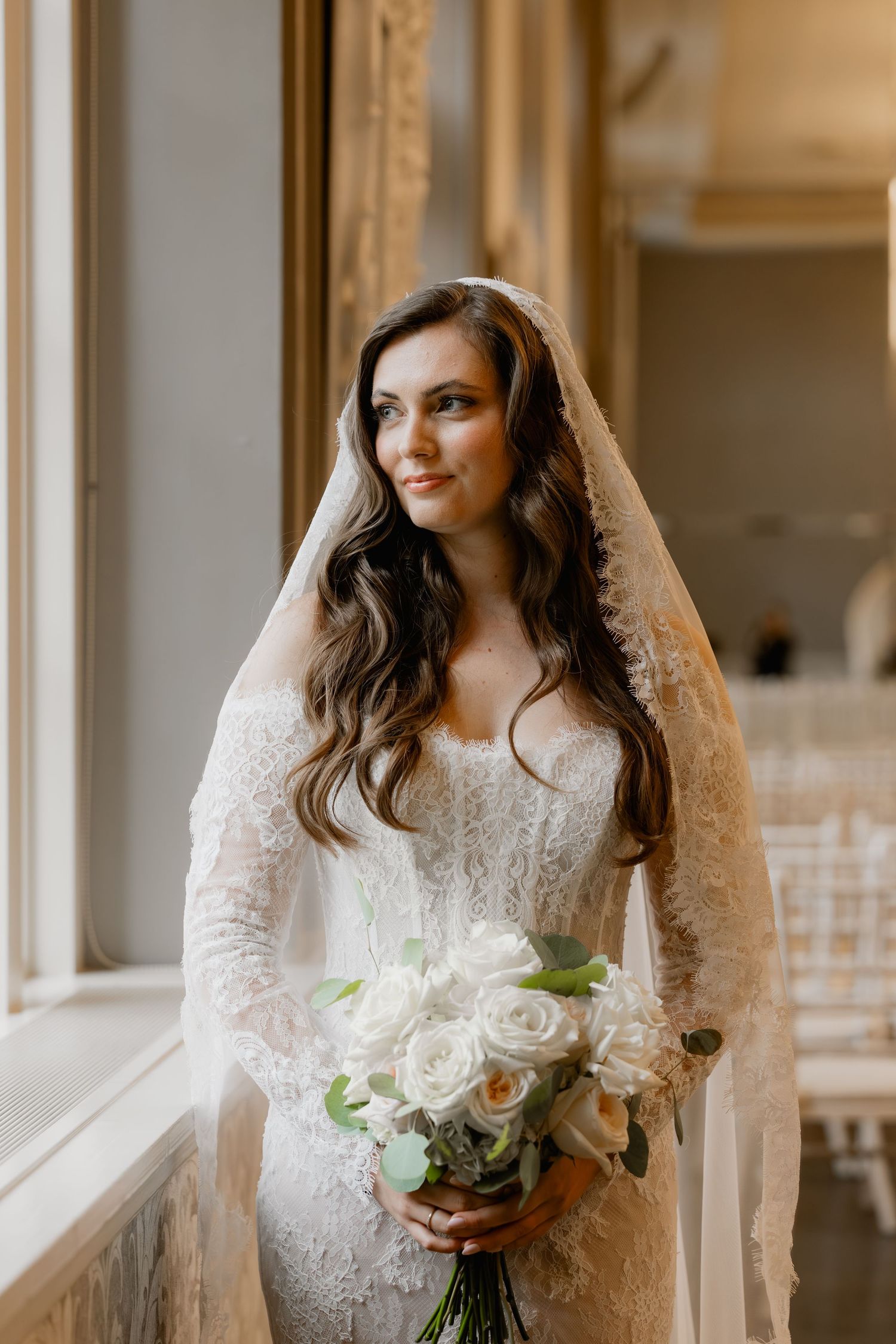 Elegant bride in lace gown and veil holding white rose bouquet standing by window in ornate venue.