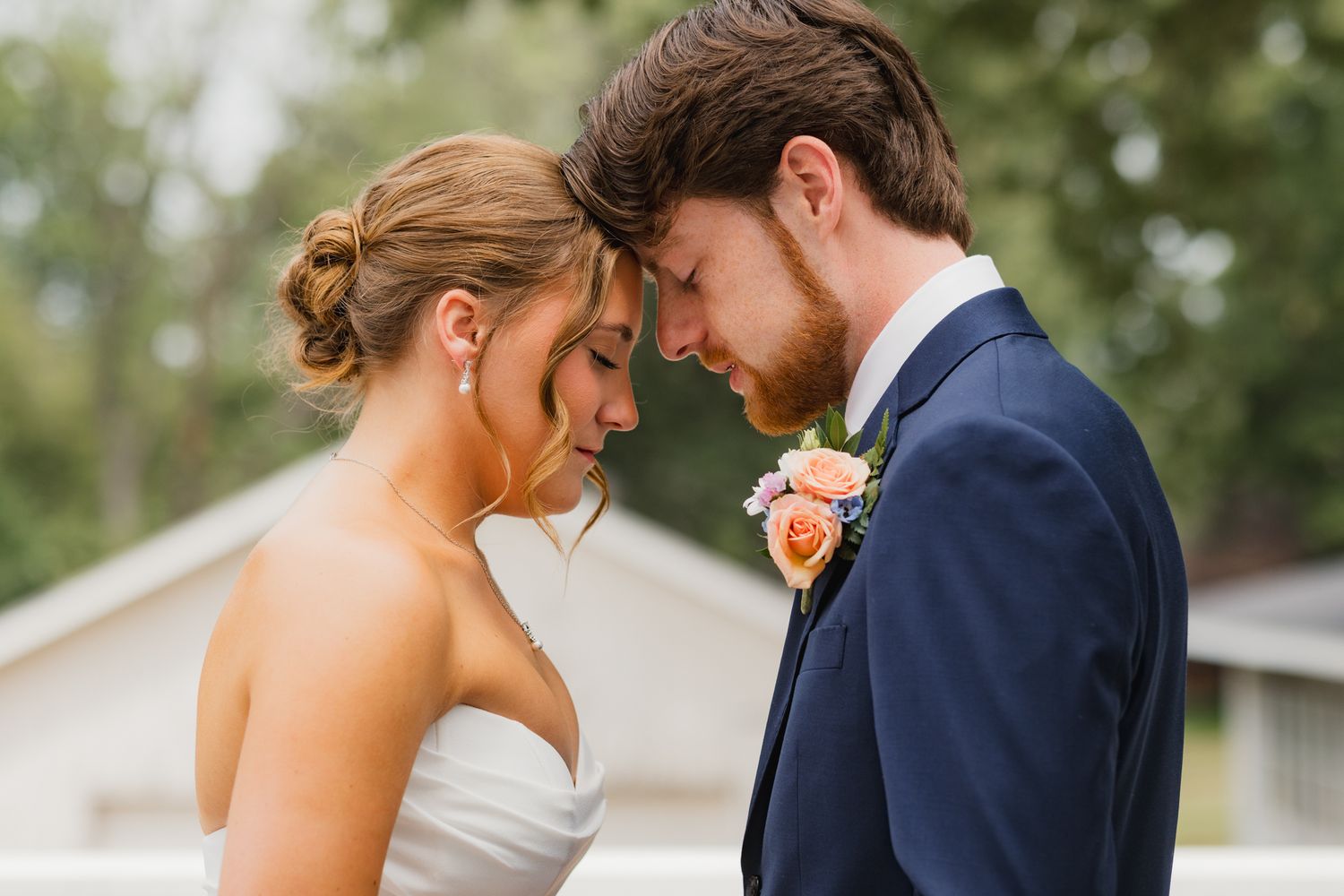 Bride and groom touching foreheads in an intimate wedding moment, wearing white dress and navy suit with boutonniere.