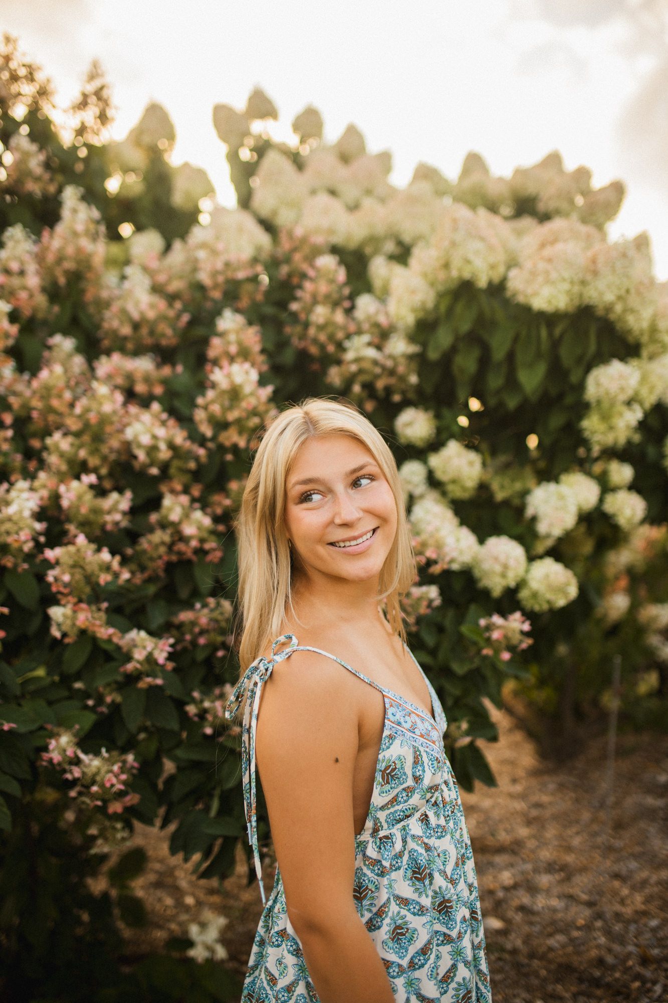 Smiling blonde woman in floral sundress posing in front of blooming hydrangea bushes in golden hour light.