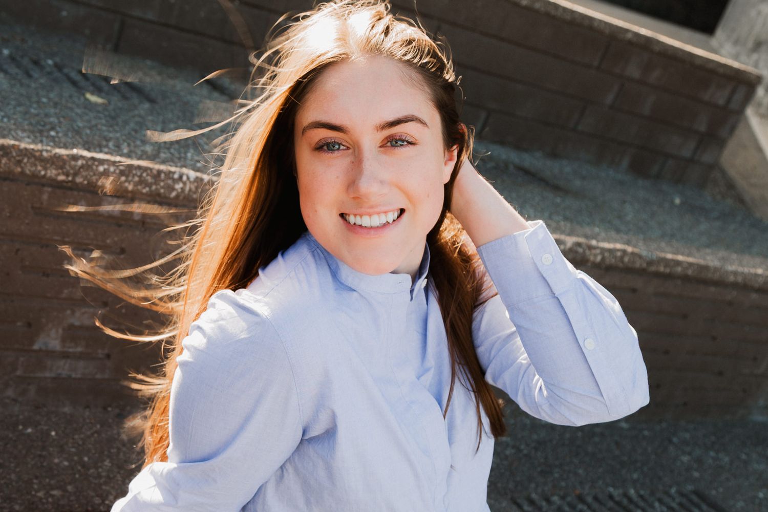 Smiling young woman with long brown hair in a light blue shirt, posing outdoors in warm sunlight.