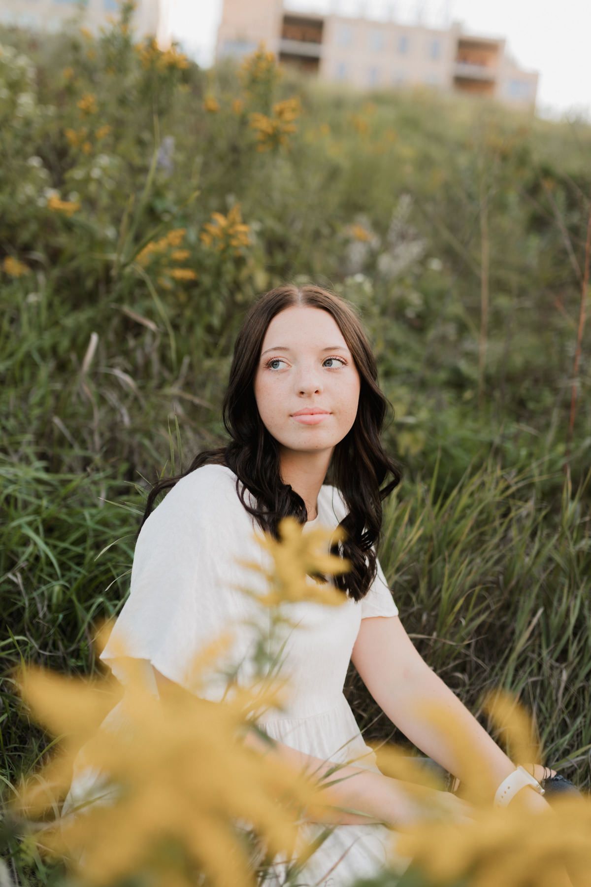 Dark-haired young woman in white dress sitting among yellow wildflowers at golden hour.