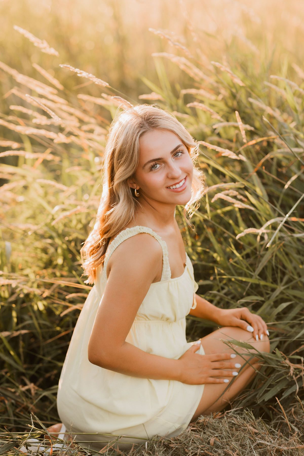 Young blonde woman in a yellow sundress sitting in a golden field of tall grass, smiling in warm sunset light.