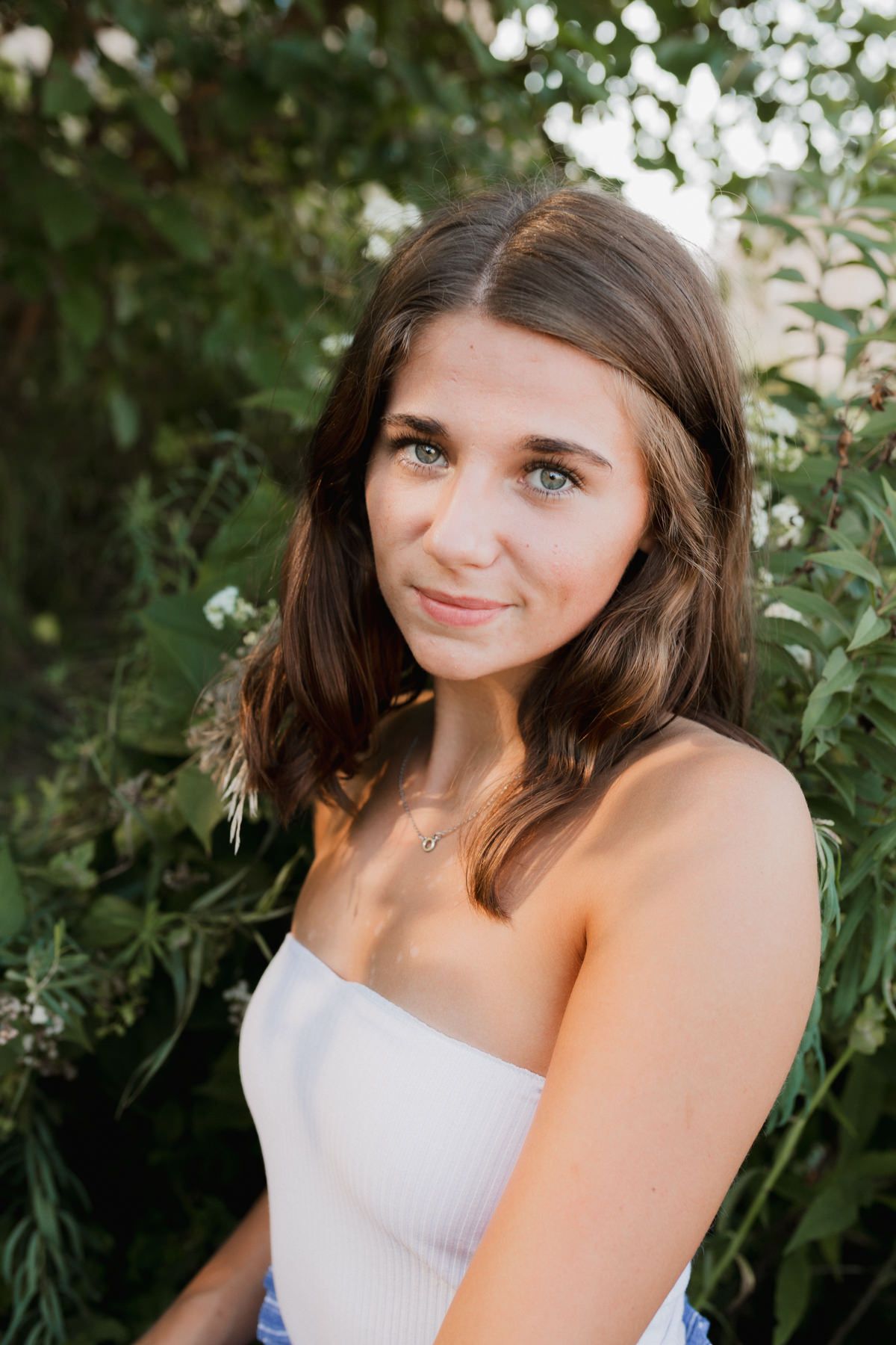 Young woman with brown hair and green eyes poses outdoors in white top surrounded by lush green foliage.