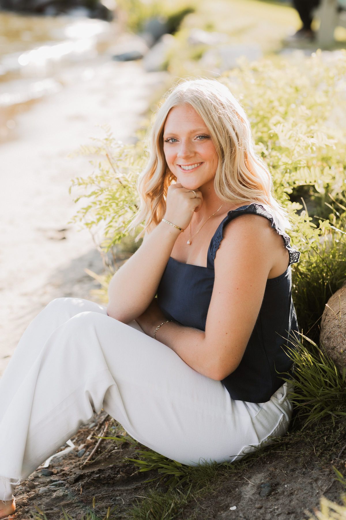 Smiling blonde teen in navy top and white pants sitting relaxed on rocks in a sunny outdoor setting.