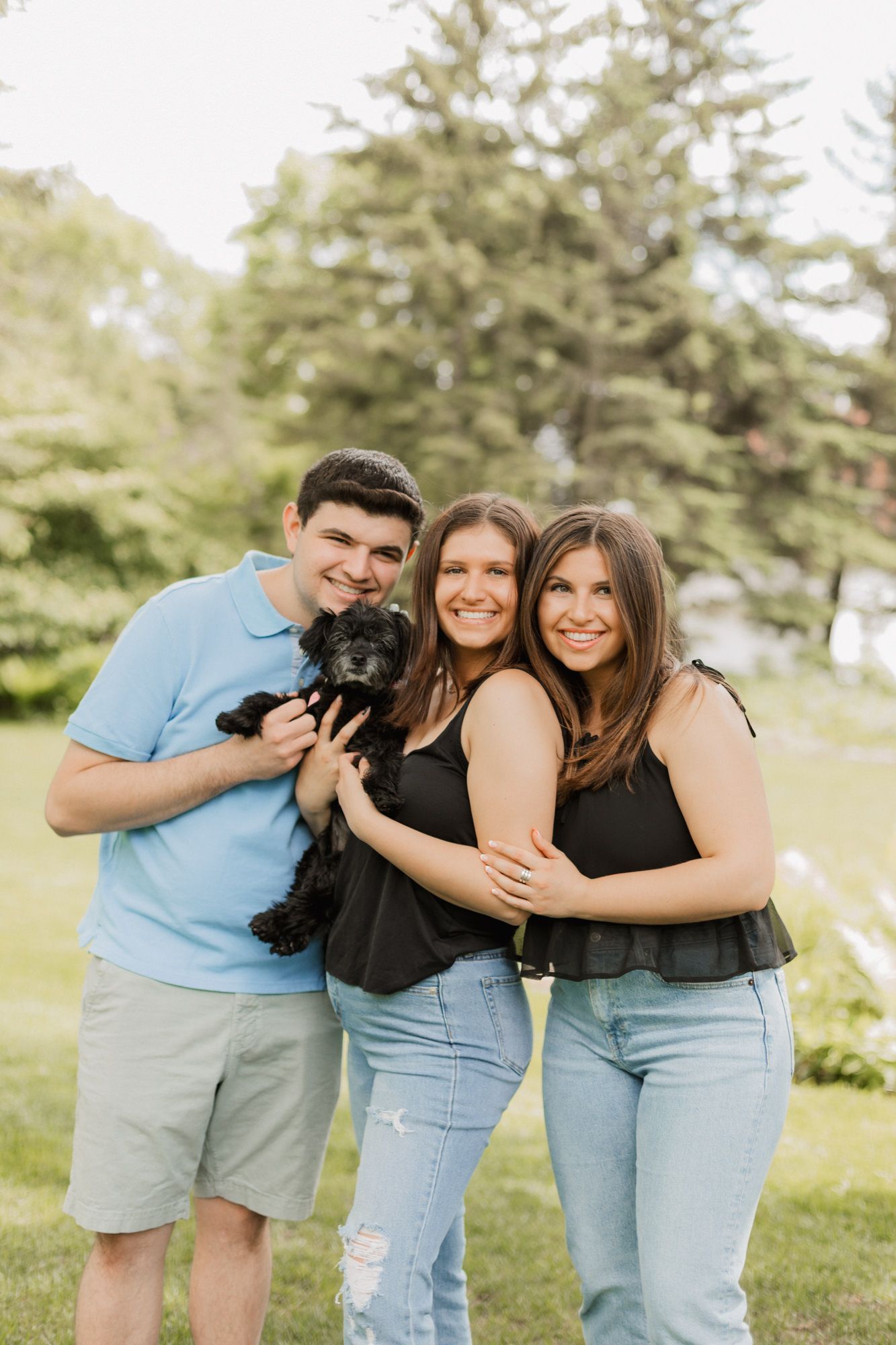 Three smiling friends pose outdoors holding a small black dog, wearing casual summer outfits on a green lawn.