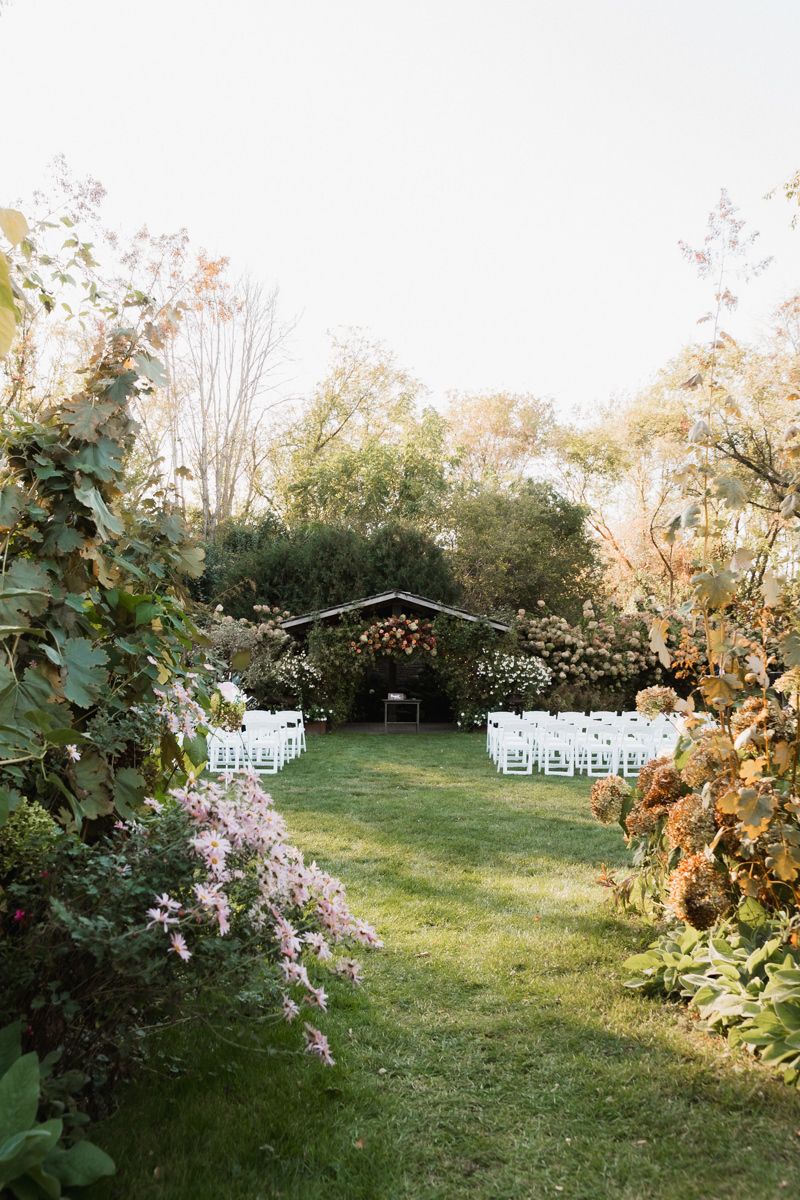 White chairs line a garden path leading to a wooden arch decorated with flowers for an outdoor wedding.