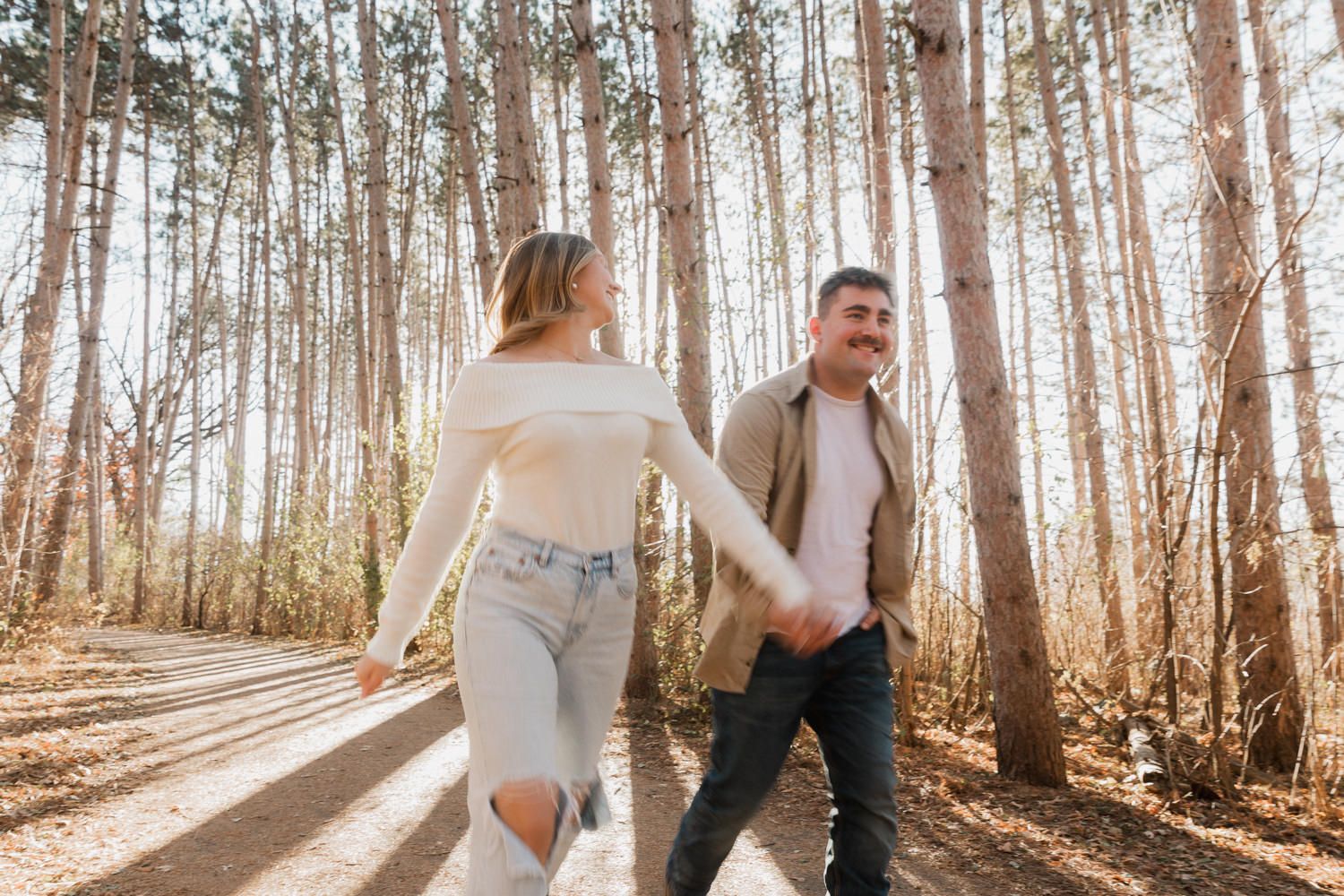 A couple walks hand in hand through a sunlit forest path lined with tall pine trees on a bright autumn day.