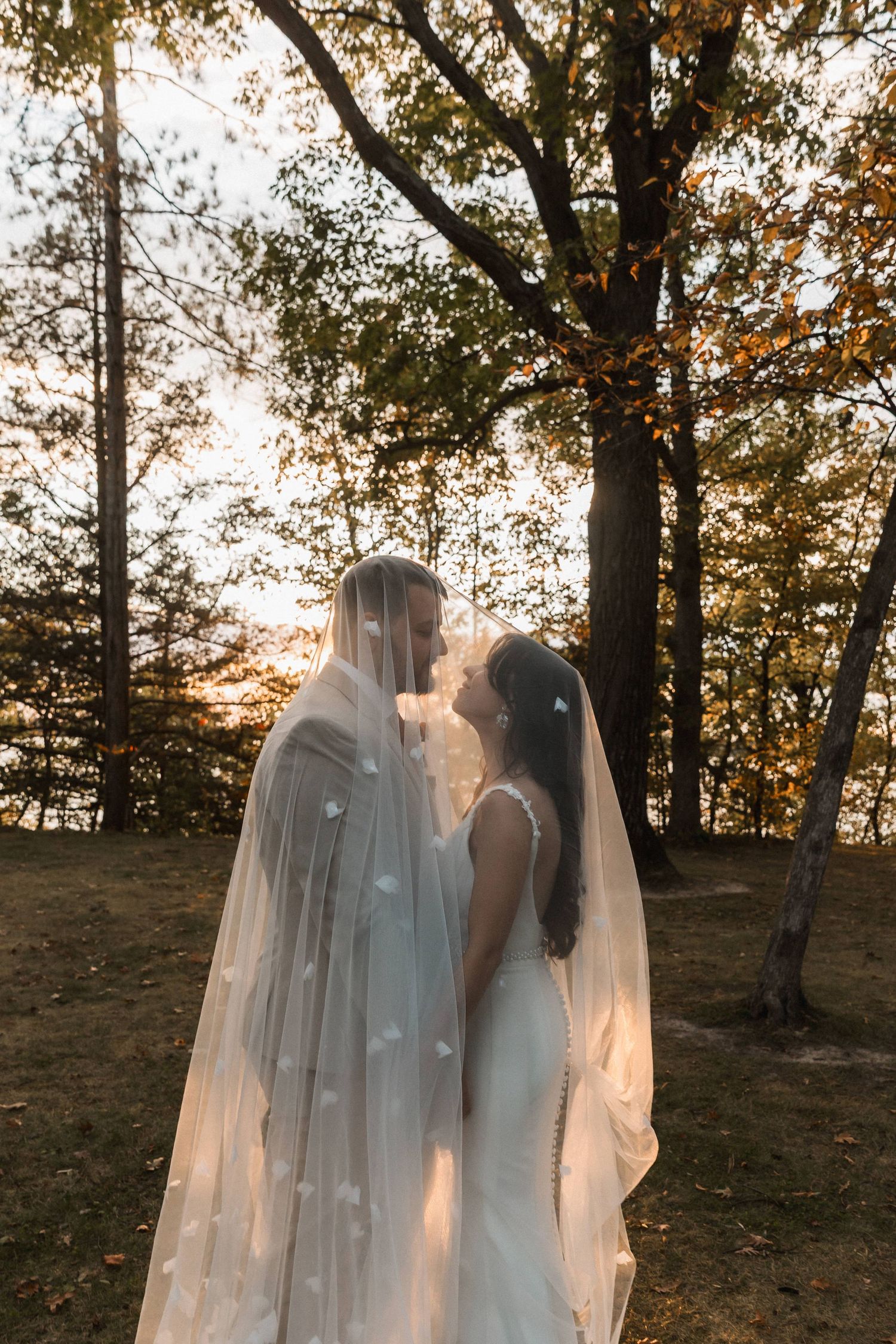 Two ethereal ghost-like figures in white veils stand together in a wooded area at sunset creating a haunting scene.