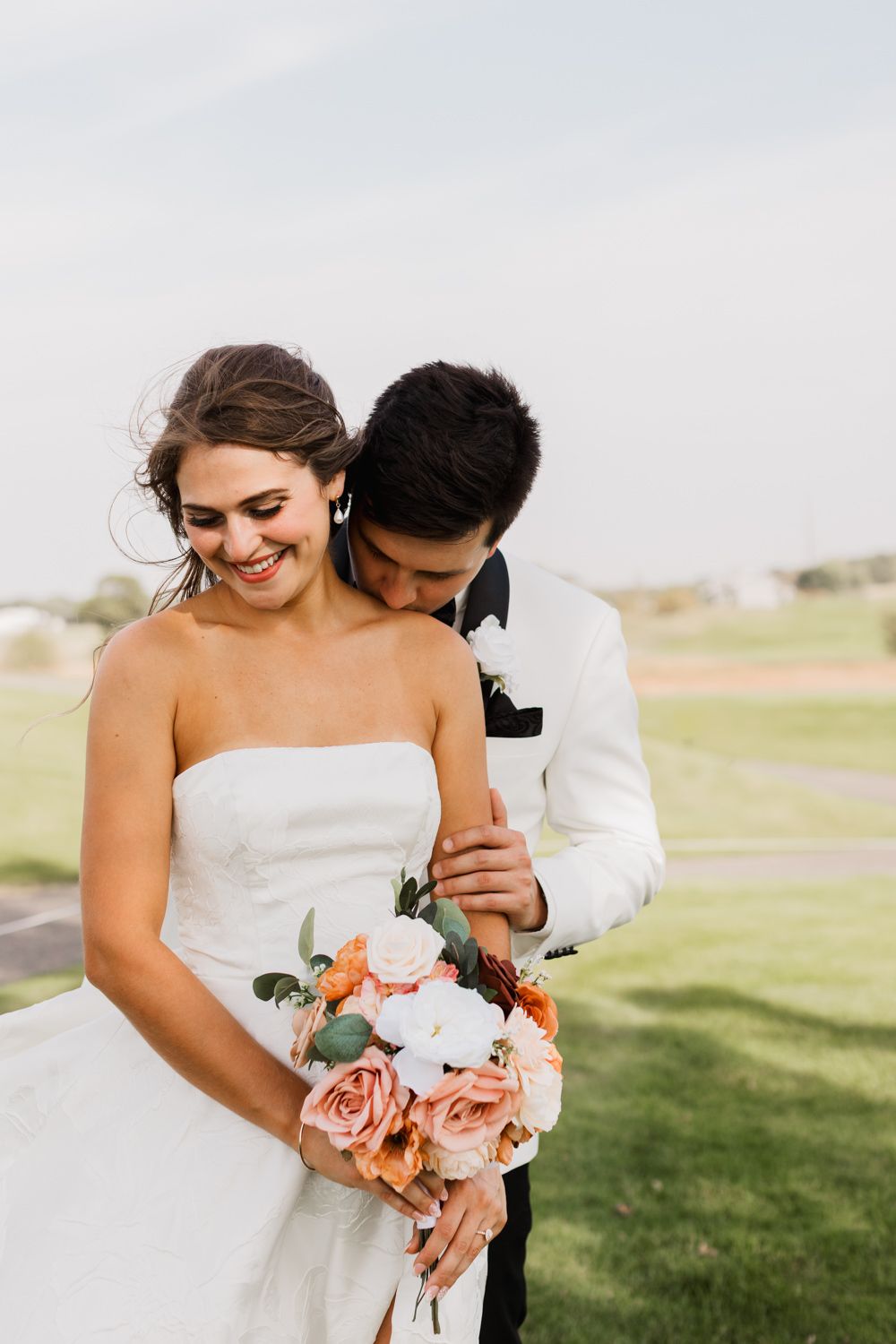 A romantic wedding moment captured outdoors with a peach and white floral bouquet against a grassy landscape.