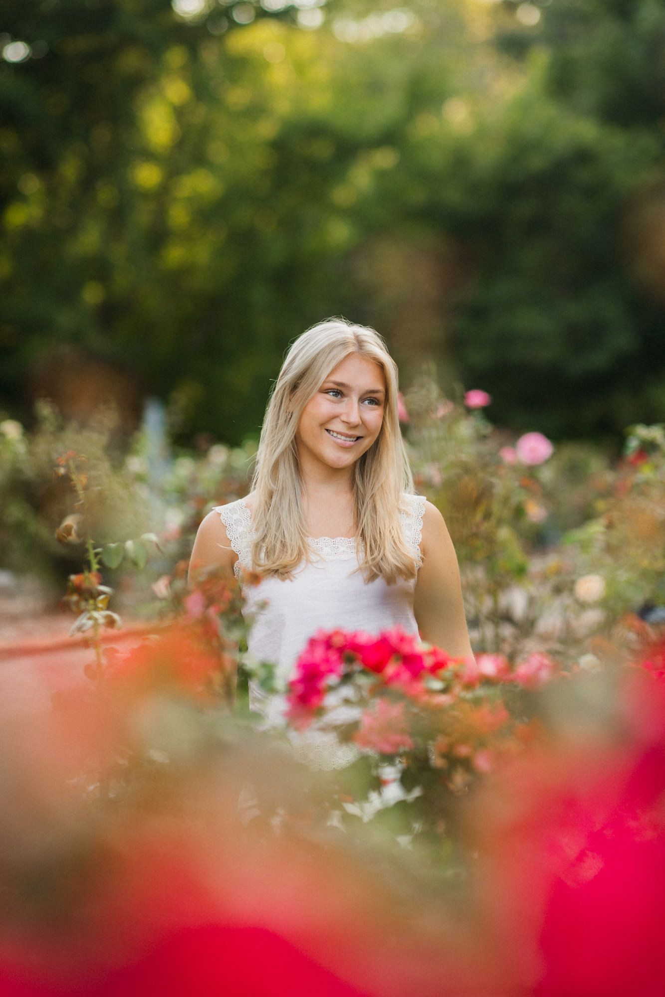Smiling blonde woman in white dress surrounded by vibrant red and pink roses in a lush garden setting.