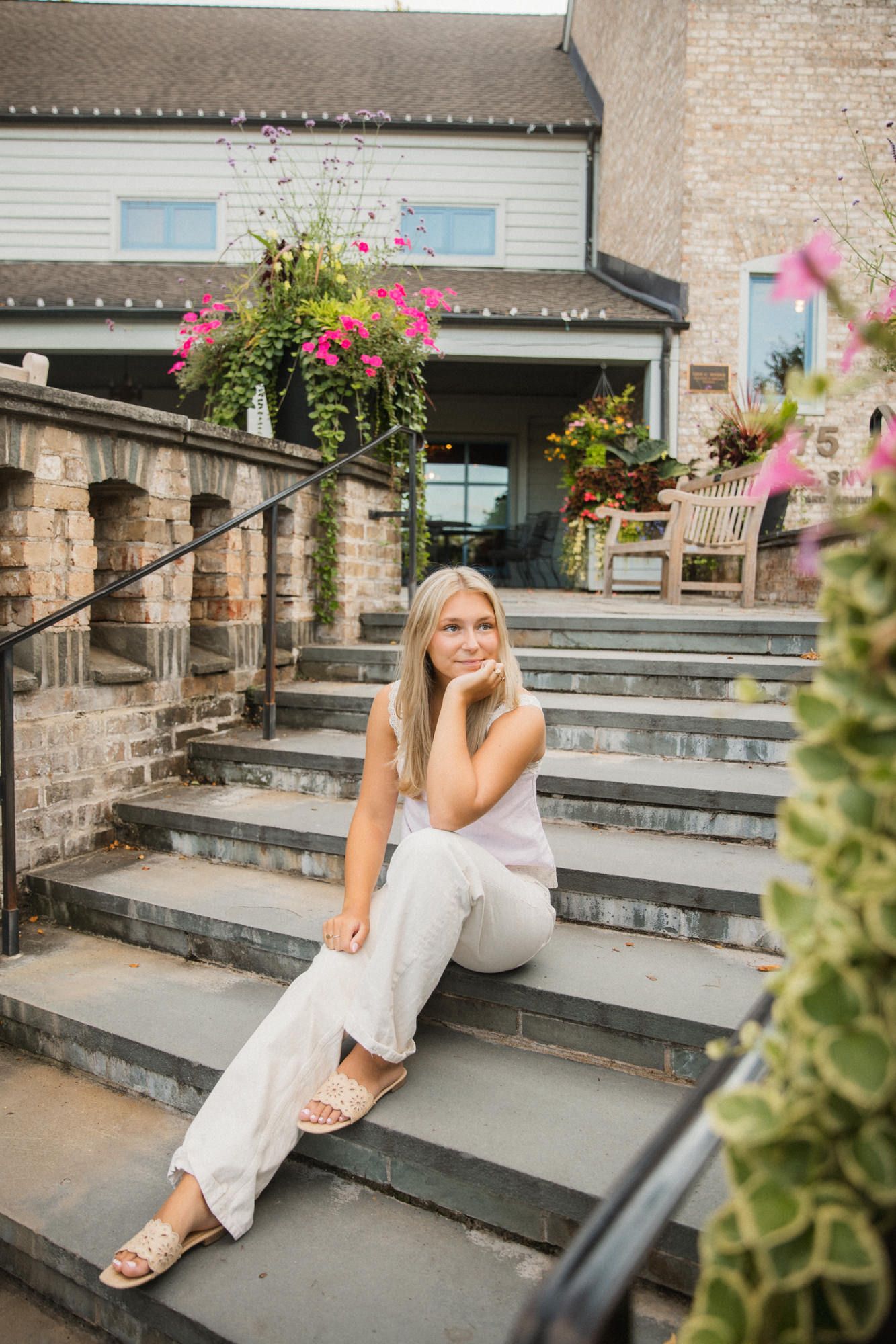 Smiling blonde woman sitting on stone steps outdoors, wearing white outfit, with pink flowers and brick building behind her.