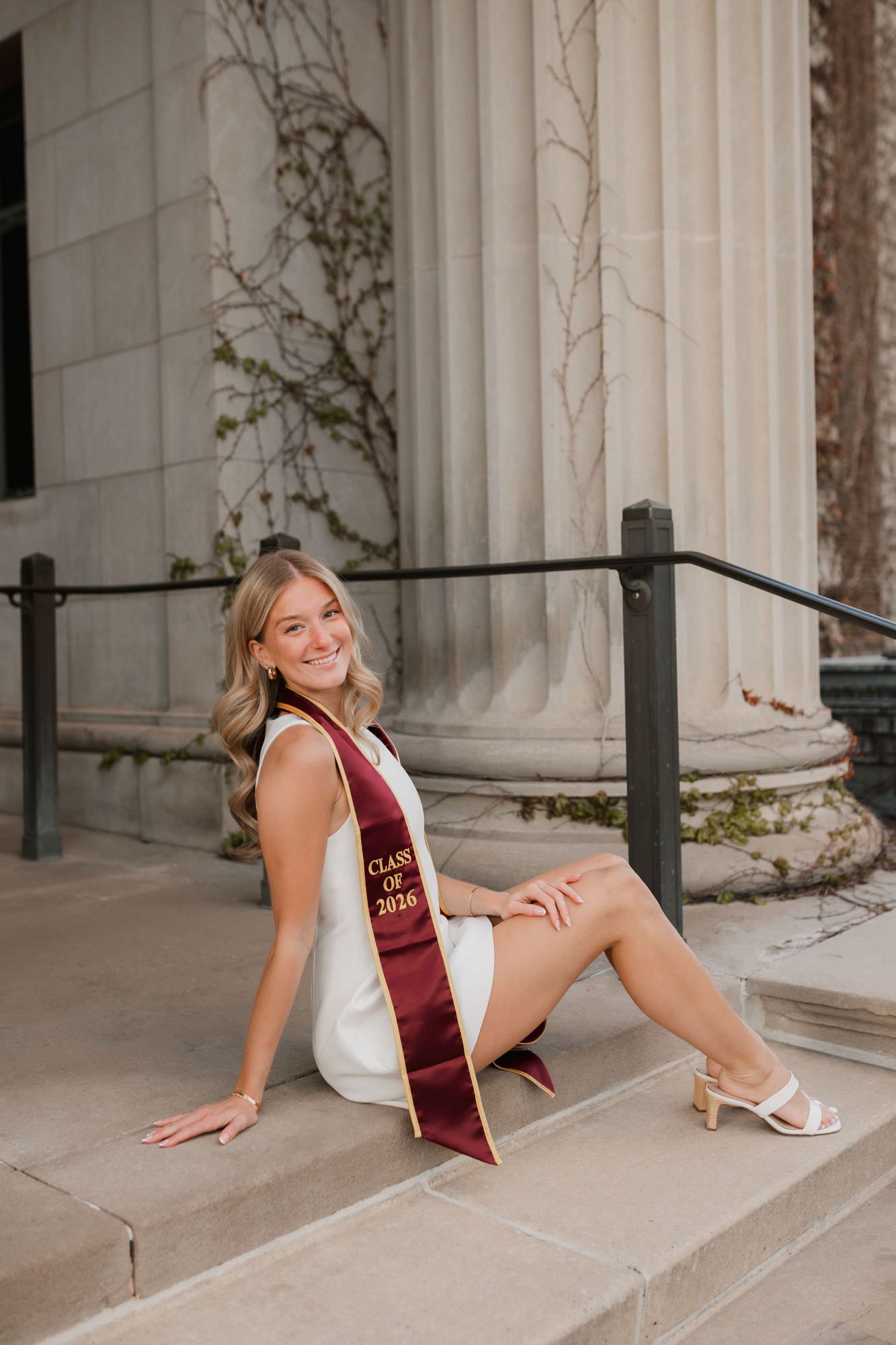 Smiling college graduate in white dress and maroon Class of 2025 stole sits on steps near classical columns.