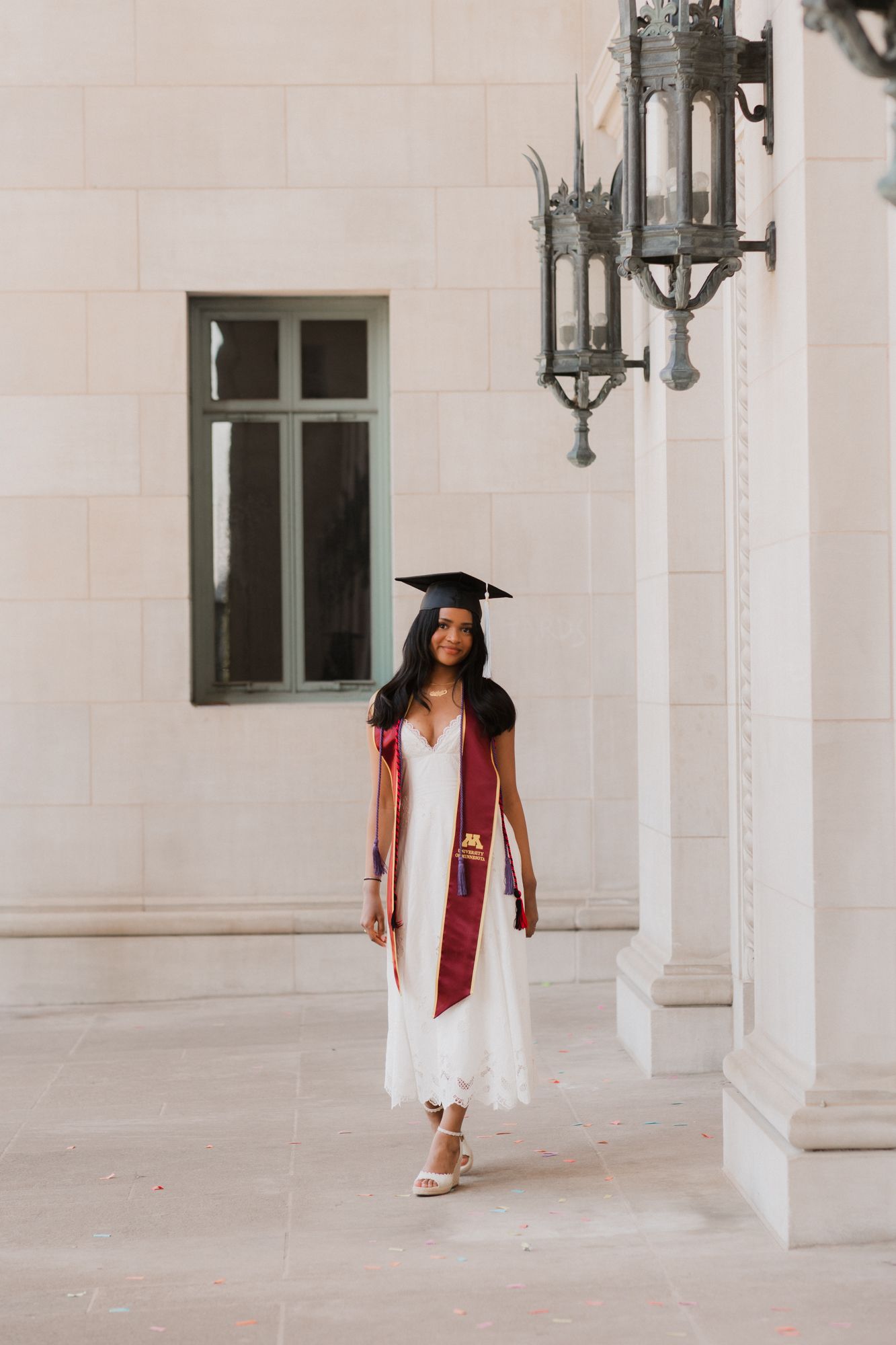 A young woman in a white dress and graduation cap and maroon stole poses outdoors near ornate columns.