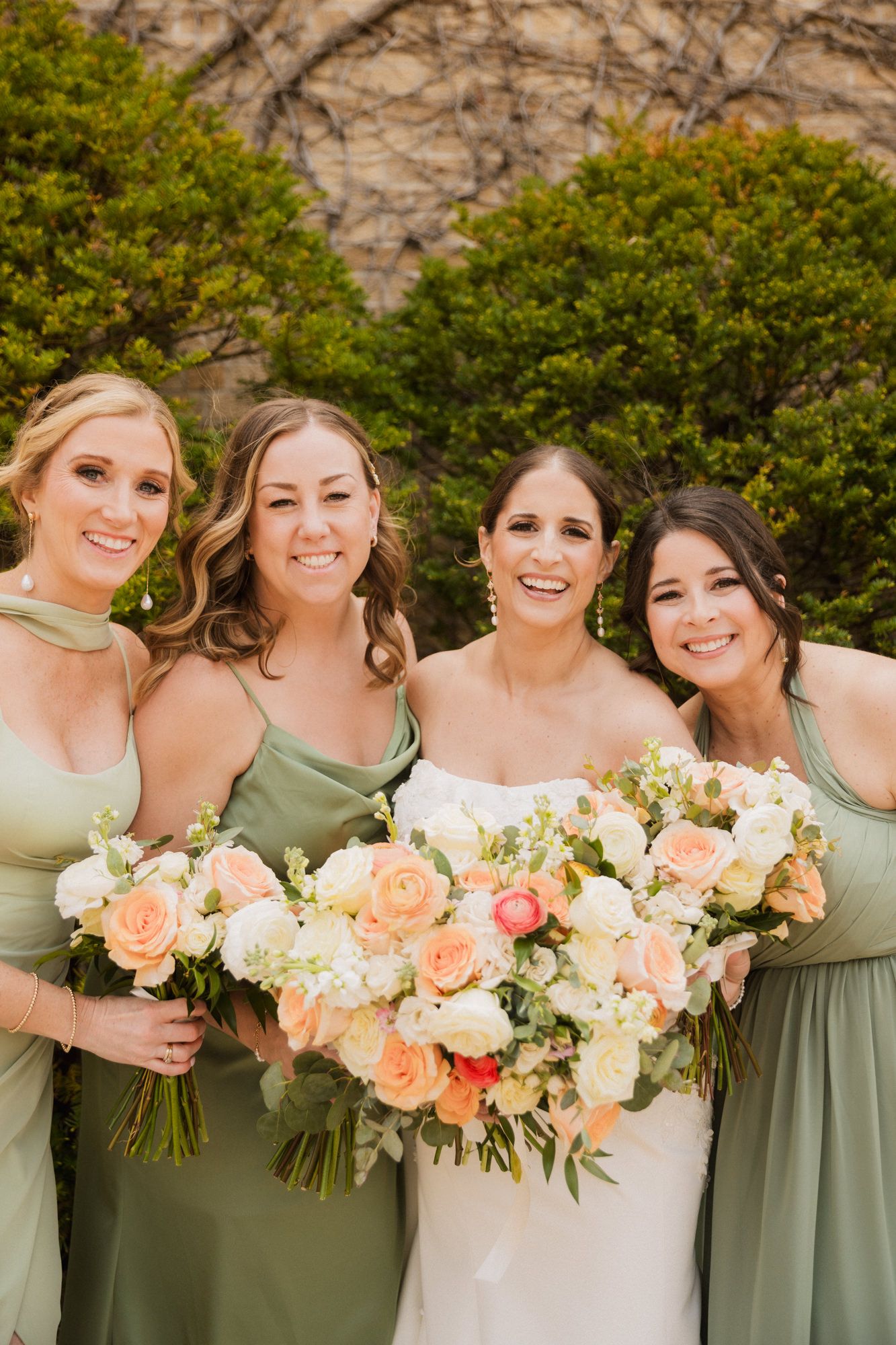 Bride in white gown with three bridesmaids in sage green dresses holding peach and white floral bouquets outdoors.