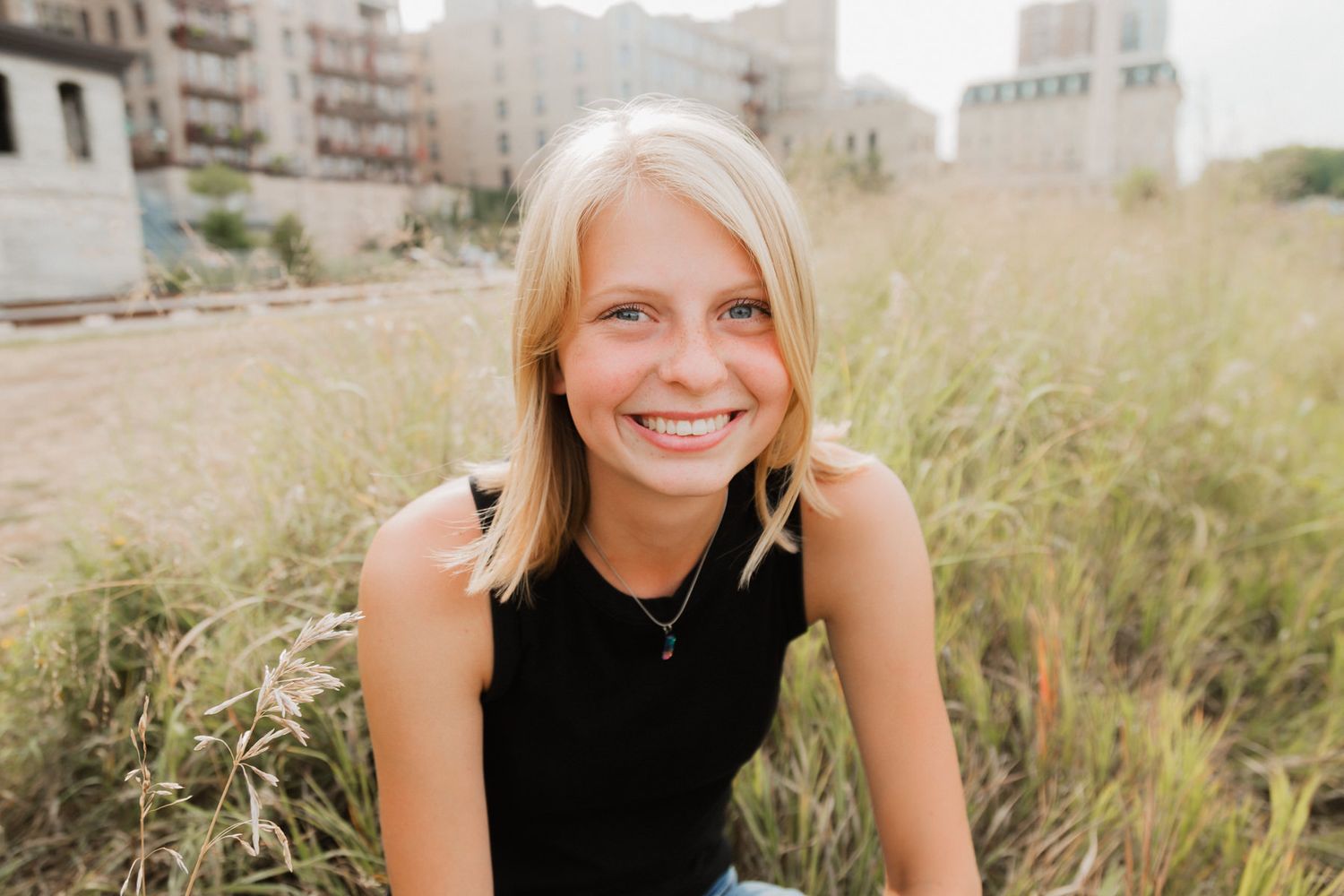 Smiling blonde teenage girl sits in tall grass with urban buildings in the background.