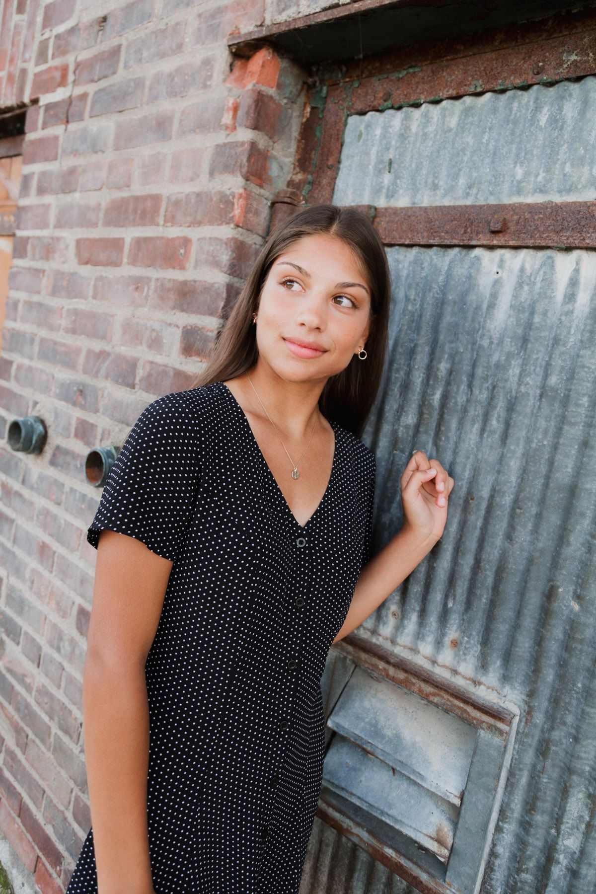 Brunette young woman in polka dot dress poses by rustic brick wall and metal door.