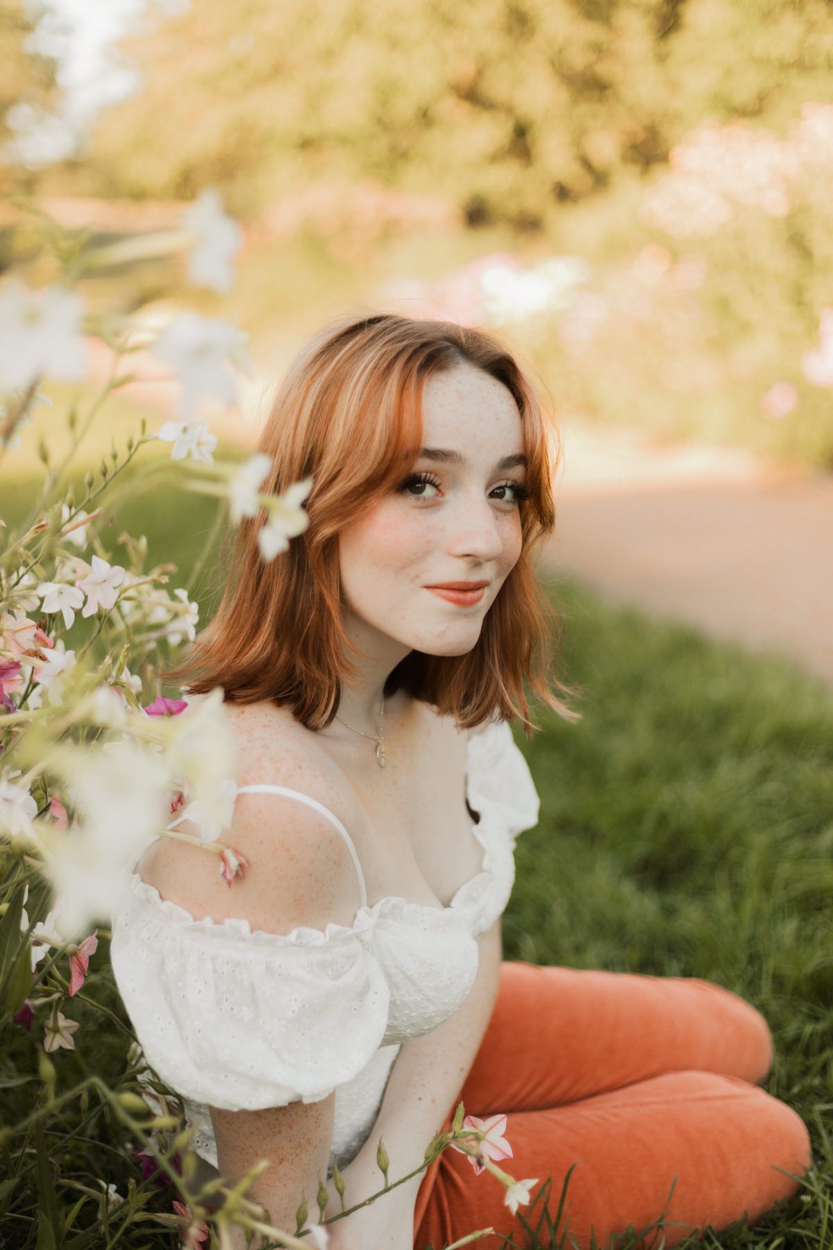 Young redhead woman in white off-shoulder top sitting in green grass surrounded by white wildflowers.