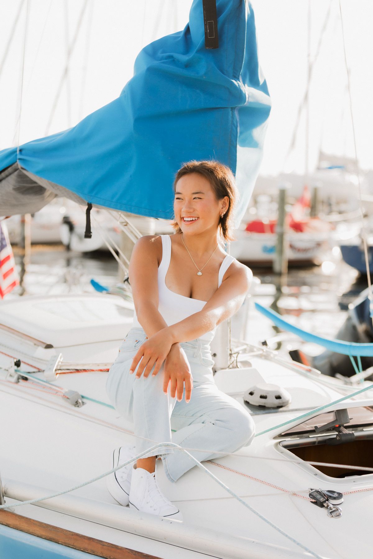 Smiling woman in white outfit sitting on sailboat deck at sunny marina with blue sail behind her.