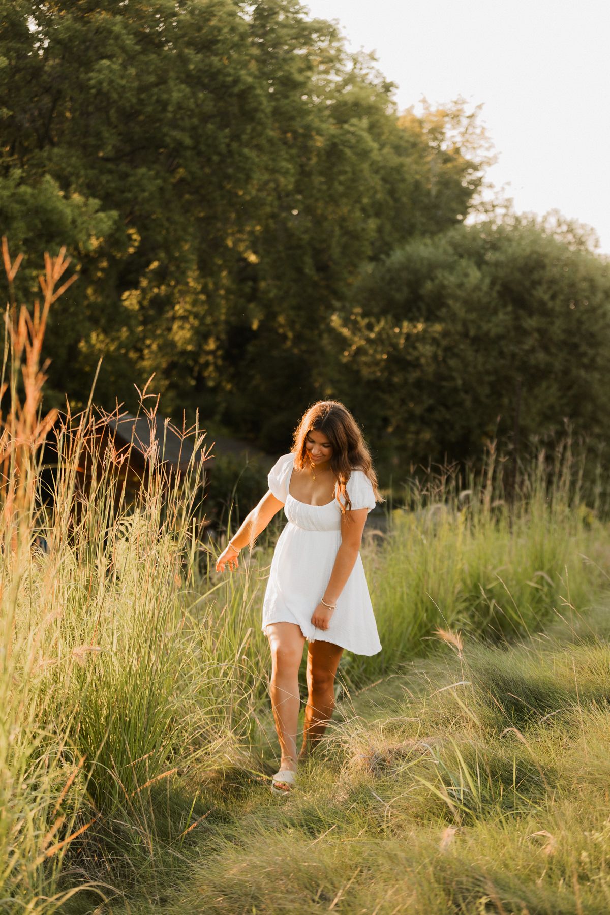Young woman in white dress walking through golden tall grass field at sunset with green trees background.