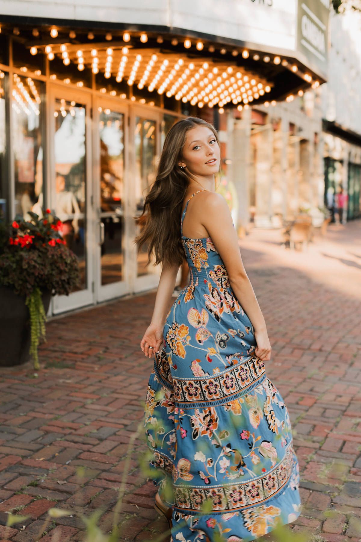 Woman twirling in blue floral maxi dress outside illuminated storefront at sunset.