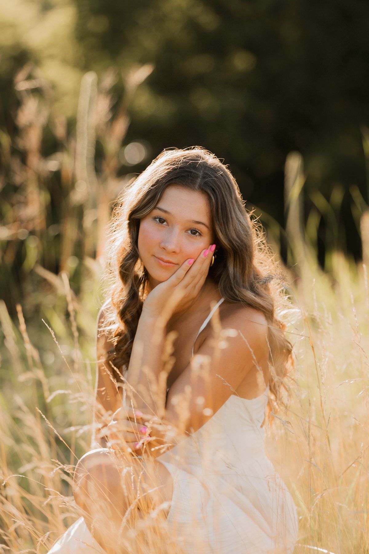 Young woman with long wavy hair sitting in golden field grass, wearing white dress, golden hour sunlight.