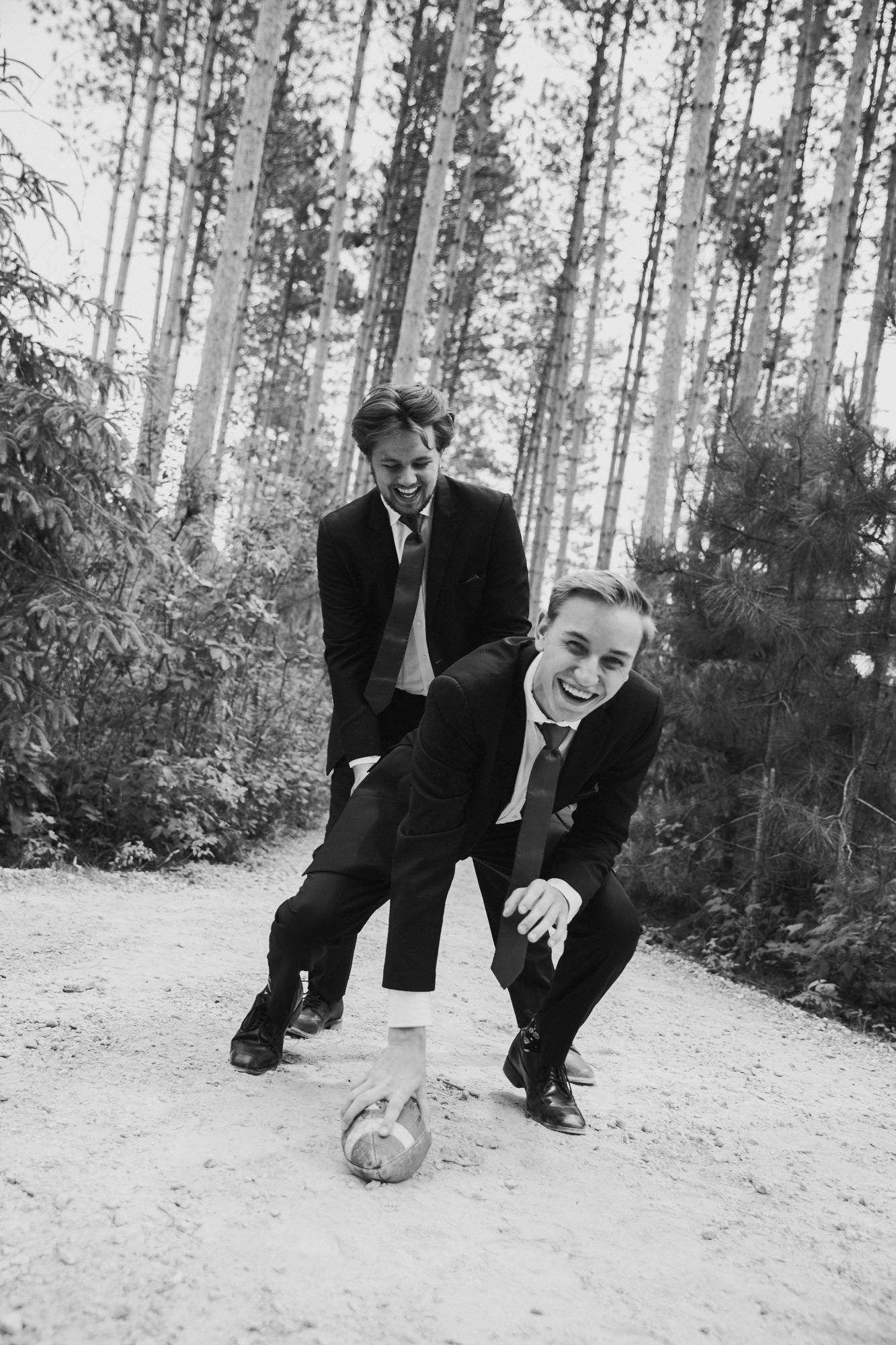 Two men in suits laughing while playing with a soccer ball on a sandy forest path in black and white.