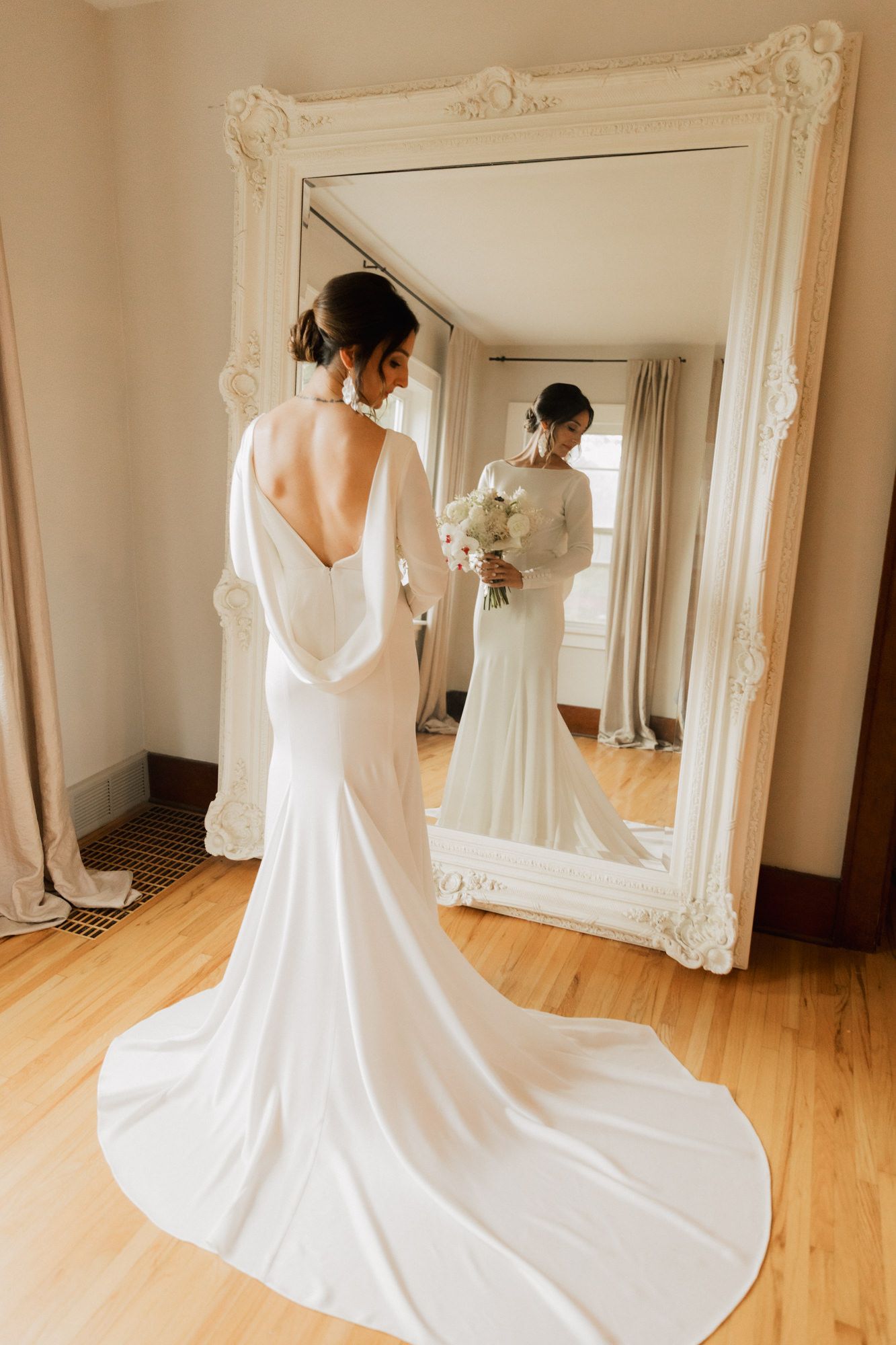 Bride in elegant white backless wedding gown with train stands before ornate mirror holding bouquet.