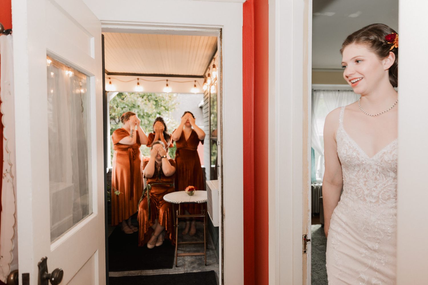 Bride in white lace gown smiles as bridesmaids in rust dresses react with excitement in doorway during wedding day reveal.