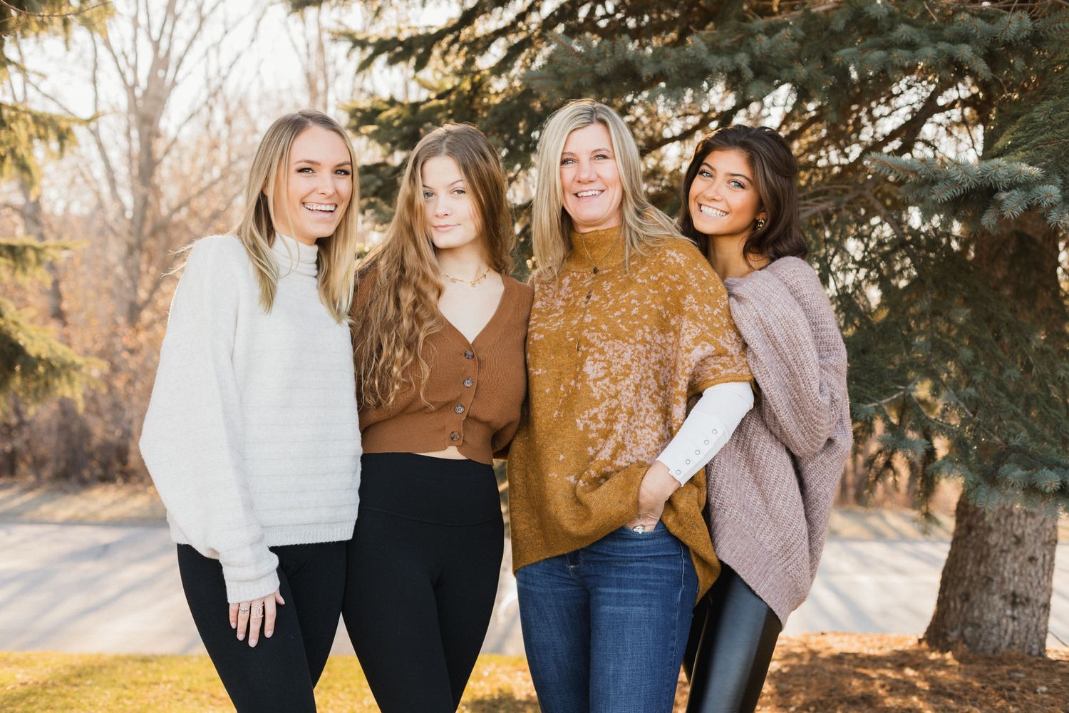 Four women smiling outdoors near a pine tree in fall, wearing cozy sweaters in neutral tones.
